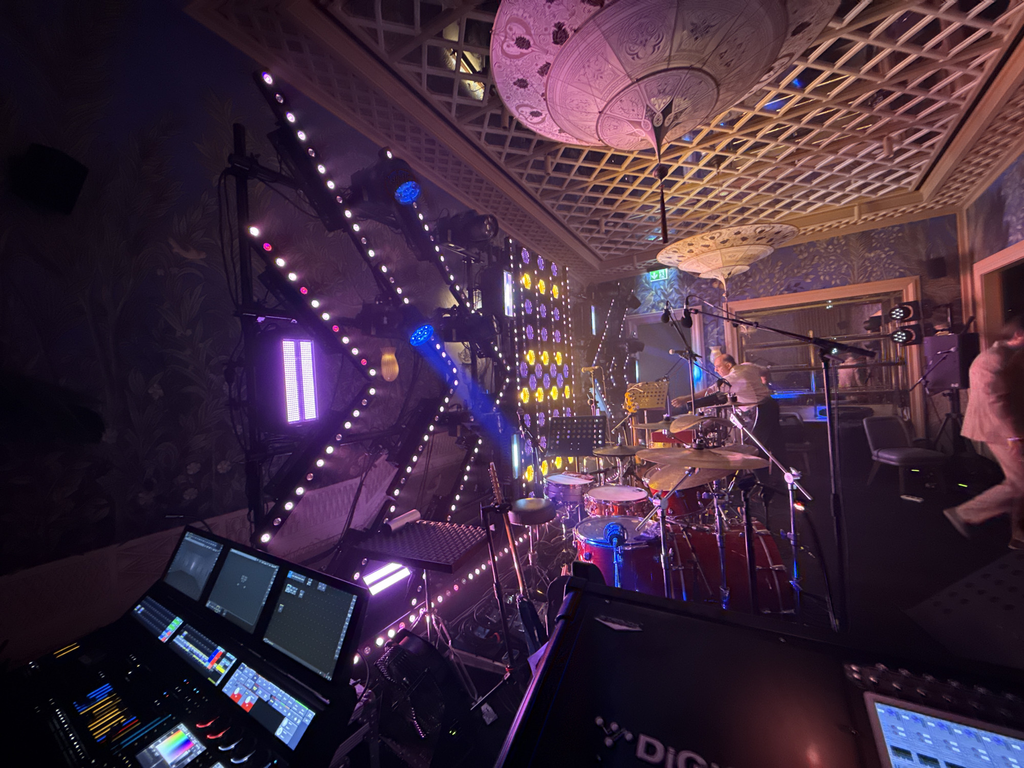 The image shows a stage setup with various lighting equipment and musical instruments. In the foreground, a large audio mixing console with multiple screens and faders is visible. To the right, a drum set is set up with cymbals and drums, some of which are red. A person is partially visible playing the drums. Behind the drum set, there are several microphones and stands.
The stage is adorned with numerous lights, including rows of LED lights and larger spotlights, casting a purple and yellow glow across the scene. The background features a wall with a patterned wallpaper and a decorative ceiling with a lattice design and two large, ornate lamps.
The overall impression is that of a live music performance setup, likely in an indoor venue. The specific location in Baku, Azerbaijan, is not identifiable from the visual cues within the image. The image captures a moment of preparation or performance on a well-lit stage.