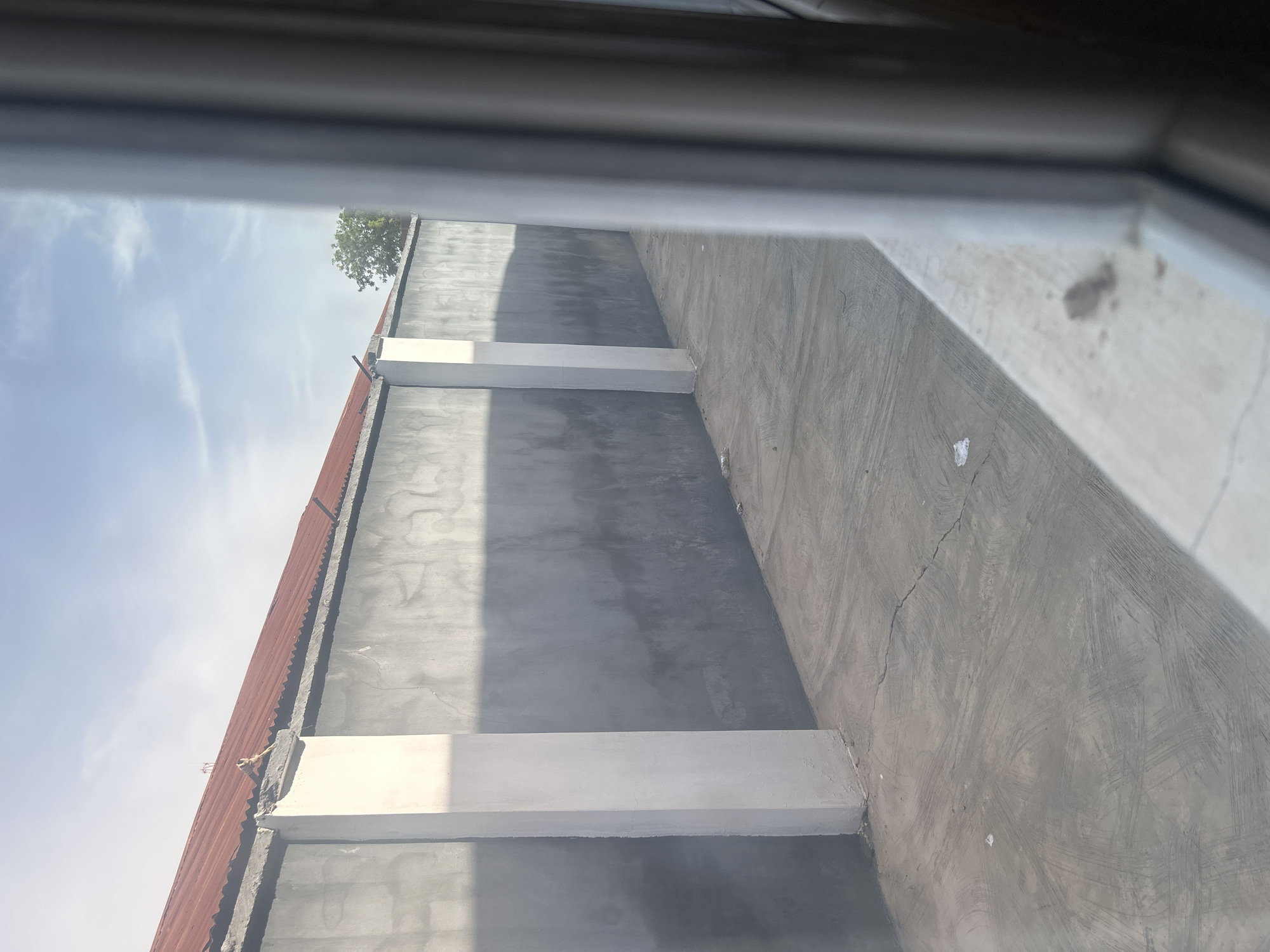 A view through a window of a narrow concrete walkway next to a gray concrete wall. The walkway has a rough texture with some cracks and debris. The wall is tall and runs along the length of the walkway, with white rectangular pillars spaced intermittently. A red corrugated metal roof is visible above the wall on the left side of the image, and a tree can be seen in the distance on the right. The scene suggests an outdoor space in a residential or commercial area, likely in Ogbomosho, Nigeria, given the typical construction and environment. The lighting indicates it is daytime, with shadows cast by the wall and pillars.