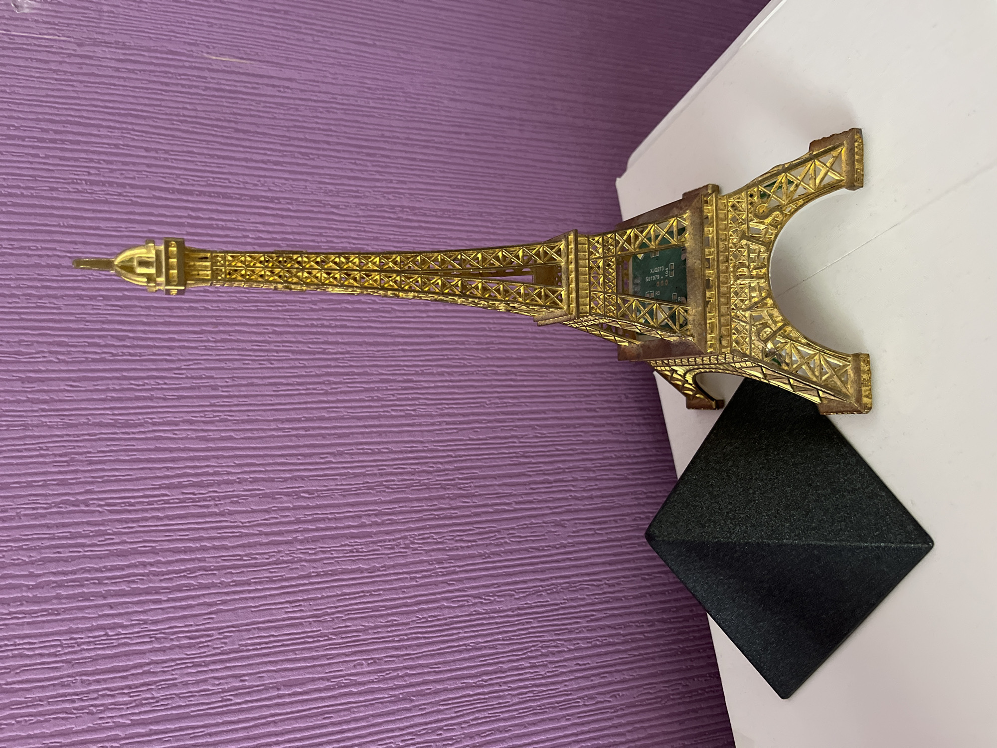 The image depicts an indoor scene featuring two decorative objects positioned on a white, reflective surface against a textured purple wall.
The primary object is a gold-colored, metallic replica of the Eiffel Tower, which stands upright on the right side of the frame. Its intricate lattice structure is visible, along with four arched base supports. Within the lower section of the tower, a green circuit board is visible, displaying text that includes "X_D2021S", "S59172A", "000", and "R3", indicating an internal electronic component. The top of the tower features a small, open-arched finial. The golden finish shows some signs of wear or patina.
To the left of the Eiffel Tower, a matte black, square-based pyramid object is positioned. Its surfaces appear to have a slightly coarse or textured finish, reflecting minimal light.
Both objects rest on a smooth, white surface that reflects light, suggesting it is a table, shelf, or desk. The surface has a subtle beveled edge visible on the far right.
The background consists of a solid purple wall with a distinct vertical textured pattern, resembling parallel, irregular grooves or striations. No people are visible, and no actions or interactions are depicted. The lighting is even and diffused. The specific location in Vratsa, Bulgaria, cannot be determined from the visual evidence provided.