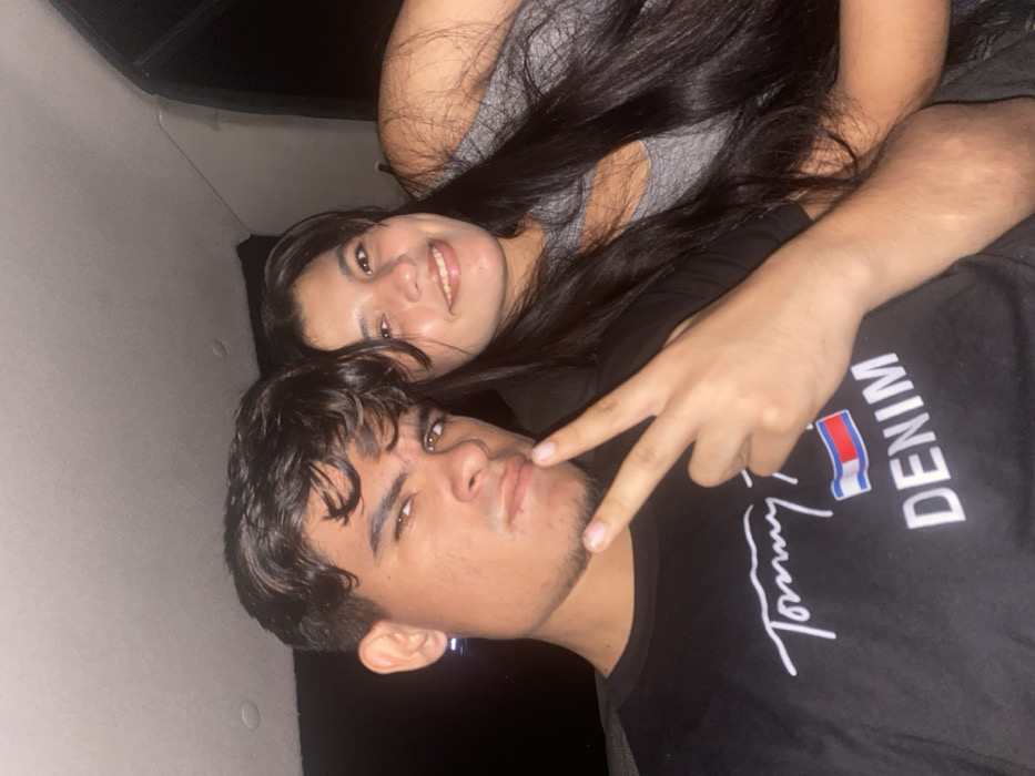 A young couple poses for a photo. The man, with dark hair and a "Tommy Denim" t-shirt, flashes a peace sign. The woman, with long dark hair, smiles from behind him. They appear to be indoors.