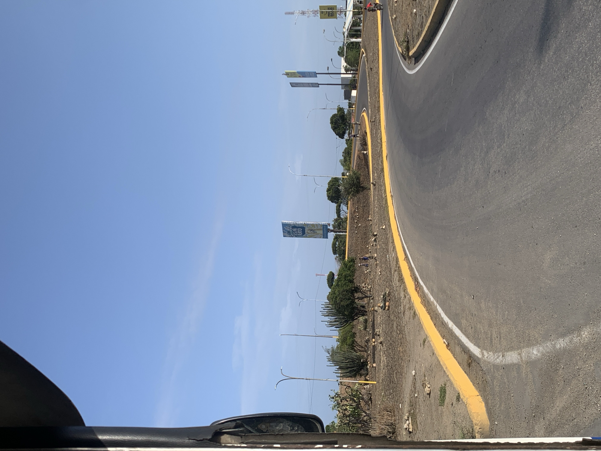 The image displays a wide asphalt road curving gently to the right, viewed from within a vehicle, with a portion of the car's interior visible in the bottom left. The sky above is a clear, bright blue, indicating good weather and bright daylight, likely midday or early afternoon.
To the left of the road, a wide, dry median or shoulder extends, characterized by sparse vegetation including bushes, cacti, and small trees, along with visible dirt and rocks. This arid or semi-arid landscape is consistent with the climate of El Valle del Espíritu Santo, Venezuela, where Margarita Island is located. Several streetlights or utility poles line this median.
Multiple large billboards are visible along the median. One prominent blue billboard in the mid-ground displays text, partially readable as "ES EL BUENO" or "ES O BUENO," suggesting a Spanish-speaking location. Another billboard further down appears to have the numbers "365." The road itself is clearly marked with distinct yellow and white lines, defining its edge and possibly a lane. In the distance, small human figures can be discerned on the far side of the road or median, and a person on a motorcycle is faintly visible further along the curving road. The scene captures a moment of travel on a well-maintained road under a clear sky.
