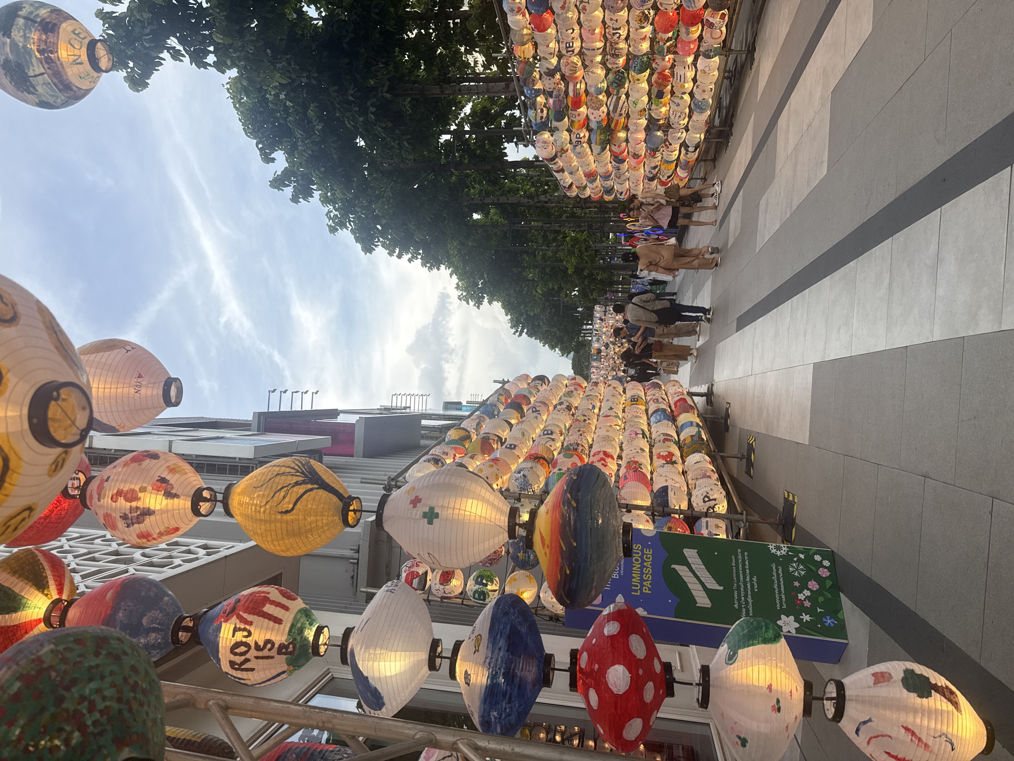 This image captures a street scene in Bangkok, Thailand, adorned with numerous colorful lanterns. The lanterns are suspended from scaffolding and are of various sizes and designs, many featuring hand-painted artwork. In the foreground, several large, intricately decorated lanterns are prominent. A blue sign with white text, reading "LUMINOUS PASSAGE" in English and Thai, is visible near the foreground lanterns, indicating a specific event or installation.
The street is paved with light-colored tiles, with darker gray stripes marking a path. On the right side of the street, a dense display of smaller lanterns stretches along a wall or structure, creating a vibrant, glowing effect. Trees with lush green foliage line the right side of the walkway, providing a natural backdrop.
In the background, people are walking along the street, some in small groups. They are dressed in casual attire. The architecture of buildings can be seen behind the lanterns, with modern elements and windows. The sky above is partly cloudy, with hints of blue sky peeking through. The overall atmosphere suggests a lively outdoor event or a decorated thoroughfare, likely in a commercial or entertainment district of Bangkok.