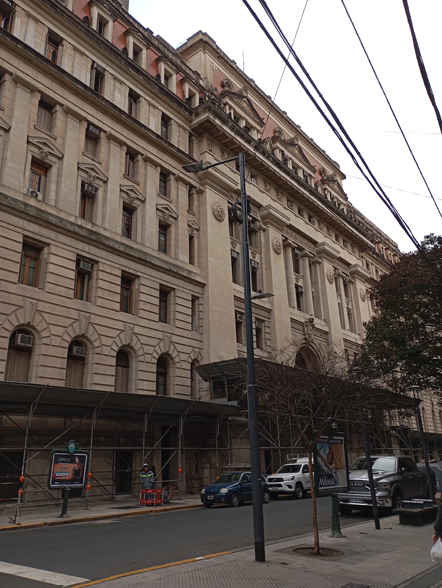 The image captures a wide street in Buenos Aires, Argentina, with a large, ornate stone building dominating the background. The building exhibits classical architectural elements such as columns, decorative moldings, and arched windows, with a red-tiled roof visible at the top. Scaffolding is erected along the lower floors of the building, suggesting ongoing construction or renovation. A street sign with the name "CAMILO" and an advertisement for a performance are mounted on a pole near the sidewalk. Several cars are parked along the street, including a blue sedan, a white SUV, and a silver pickup truck. A person in a work vest stands near the scaffolding, and another person is partially visible in the lower right corner carrying a white plastic bag. Overhead, a network of electrical wires crisscrosses the sky. The sidewalk is paved with interlocking concrete tiles, and a tree with bare branches stands near the curb. The overall scene suggests a bustling urban environment in Buenos Aires.