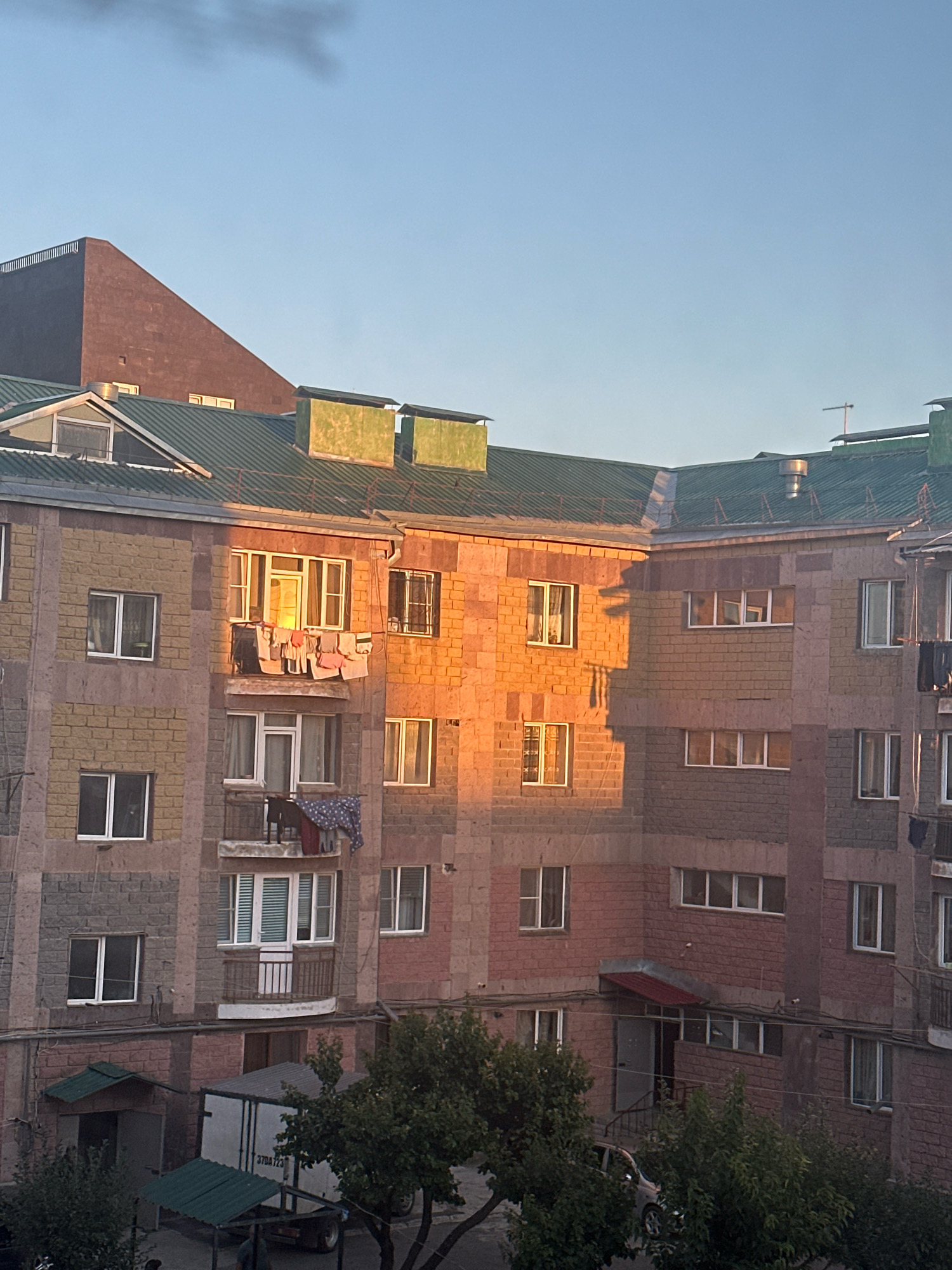 The image depicts a multi-story apartment building in Gyumri, Armenia, bathed in the warm glow of sunset. The building features a facade constructed with a mix of light beige and pinkish-brown stones. Various windows, some with white frames and others with darker ones, punctuate the structure. Laundry hangs from a clothesline on a balcony on the third floor, with items of various colors drying in the light. Below, on the ground floor, are entrances to two small structures with green corrugated roofs. A white truck with black lettering and numbers on its side is parked in front of one of the entrances. Several trees with green foliage are visible in the foreground, partially obscuring the lower part of the building and the surrounding area. The sky above is clear and light blue.