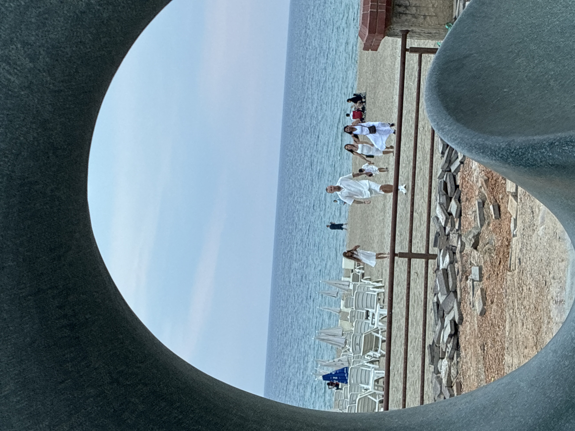 The image depicts a beach scene in Alanya, Turkey, viewed through a large, curved, gray structure, possibly part of a sculpture or architectural feature. The foreground shows a sandy area with scattered paving stones and a brown metal railing. Beyond the railing is a beach with rows of empty white lounge chairs and closed umbrellas. Several people are visible on the sand near the water. In the middle ground, a group of individuals in white clothing walks across the beach towards the viewer, with a young child in a white dress slightly ahead of them. A man in a white short-sleeved shirt and shorts walks in the foreground of this group, with two women and a small child behind him. In the background, the blue sea extends to the horizon under a pale sky. To the far right, a brick structure with a concrete column is visible.