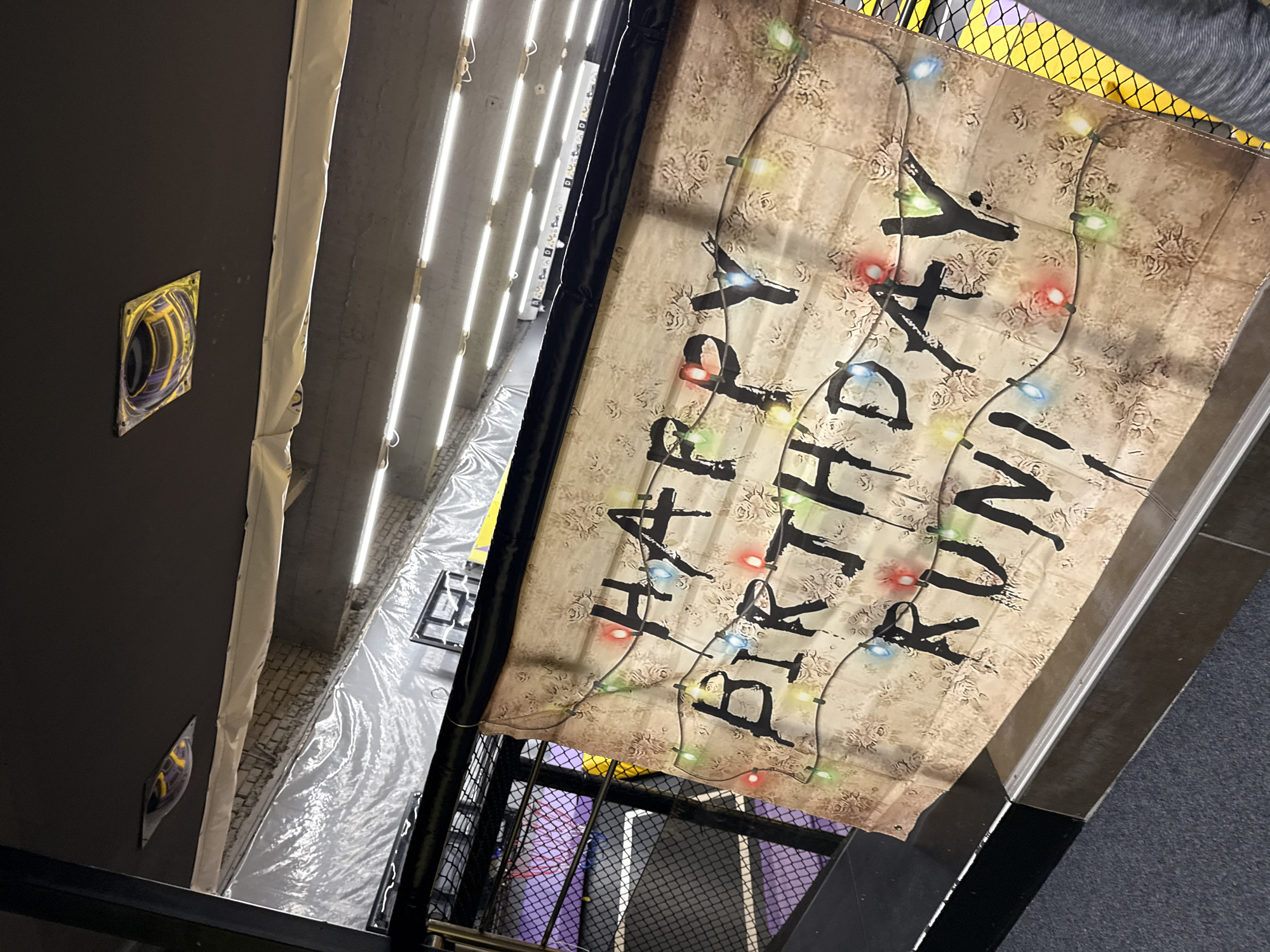 The image captures a "Happy Birthday Irwin" banner decorated with illustrations of string lights, hanging in an interior space, possibly a building in Shumen, Bulgaria. The surroundings include a metal structure with a cage-like design, suggesting a staircase or an industrial setting. The background is a mix of materials, including gray walls, bright fluorescent lighting fixtures running parallel with the ceiling, with a plastic tarp covering the floor. A few electrical outlets are visible on the wall to the left. The overall perspective is from a high angle, looking down into the space.