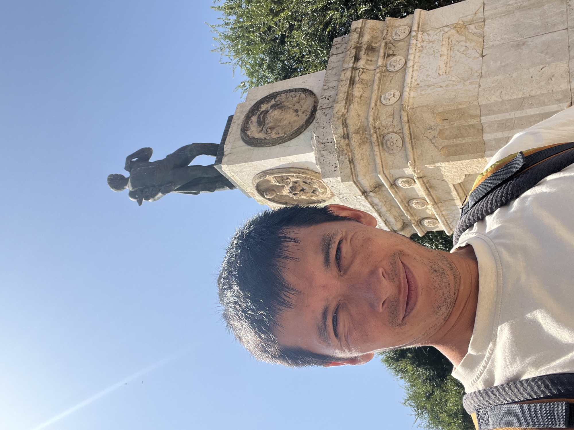 A man is taking a selfie in front of a monument in Tarragona, Spain. The man is in the foreground, smiling at the camera. He is wearing a white t-shirt and a backpack. Behind him, a statue of a man stands on a pedestal. The monument is made of stone and has intricate carvings. Trees and a blue sky are visible in the background. The sun is shining brightly, casting a lens flare on the left side of the image. The statue appears to be the Monument to Isaac Albéniz, located in the Plaça deagoza.