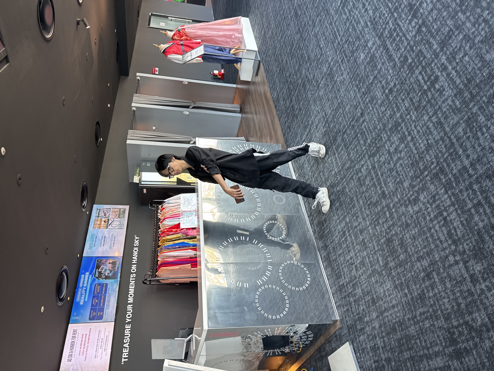 A young man wearing glasses, a black shirt, and black pants walks past a mirrored counter decorated with white circular patterns. He is holding a mobile phone in his right hand. In the background, a rack of colorful traditional Vietnamese Ao Dai dresses is visible, along with mannequins dressed in red and pink garments. Above the counter, a digital display shows information about "Ao Dai & Hanbok for Rent," "Unique Reality Games," and "Photo Service." The environment appears to be an indoor tourist attraction or souvenir shop.