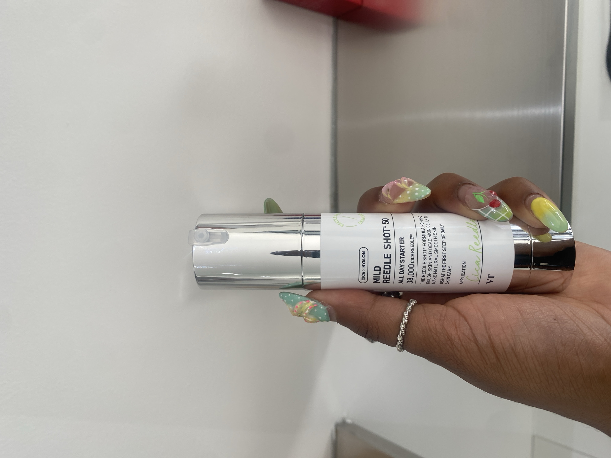 A close-up, slightly rotated shot shows a person's hand holding a silver cosmetic product bottle indoors. The person has dark skin and elaborate nail art on their fingernails, featuring green, yellow, pink, and white designs including cherries, polka dots, checkerboard patterns, and floral elements. A silver-colored twisted ring is visible on their ring finger.
The cylindrical bottle has a pump dispenser and a white label with text in black and green. Visible text includes: "CICA × HYALON", "MILD REEDLE SHOT® 50", "ALL DAY STARTER", "38,000 CICA REEDLE™", "THE REEDLE SHOT FORMULA REFINES ROUGH SKIN AND DEAD SKIN CELLS TO MAKE NATURAL SMOOTH SKIN", "USE AT THE FIRST STEP OF DAILY SKIN CARE", "APPLICATION", "Cica Reedle" (in a green cursive font), and the brand "VT". Other partially visible text includes "REEDLES" and "SKINCA". The background is a plain light-colored wall with a vertical metallic strip on the right, suggesting an indoor setting.