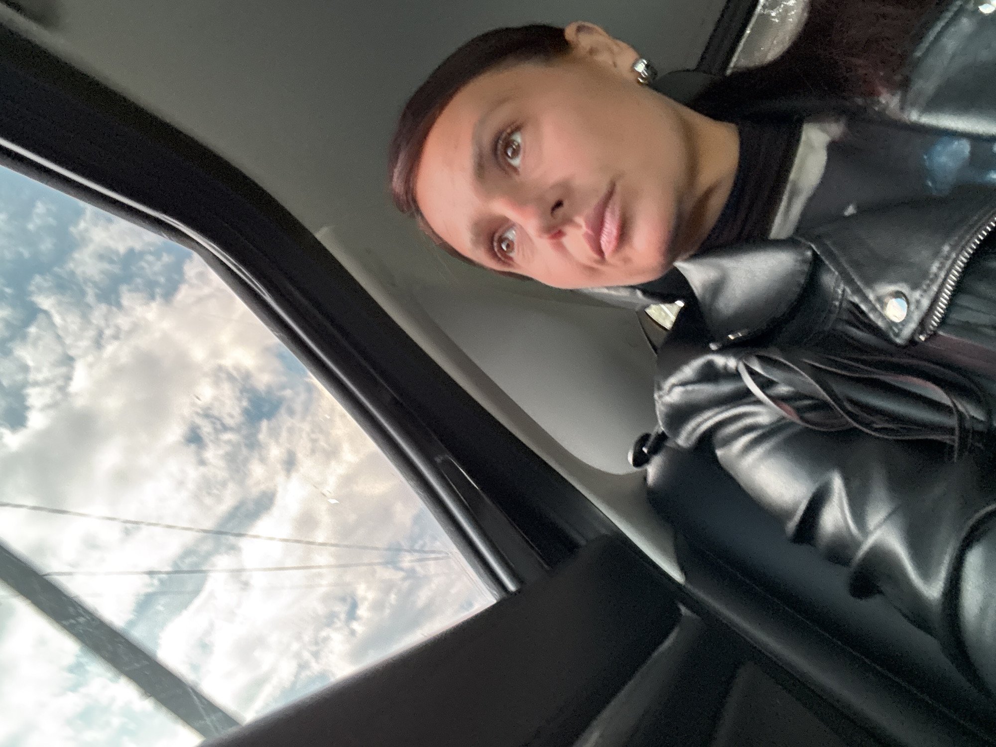 A woman is in a car, wearing a black leather jacket and a black shirt. She is looking slightly upward and to the left. Outside the car window, a cloudy sky is visible. It is not possible to determine the specific location within the city of Chaikovsky, Russia, based on the given image.