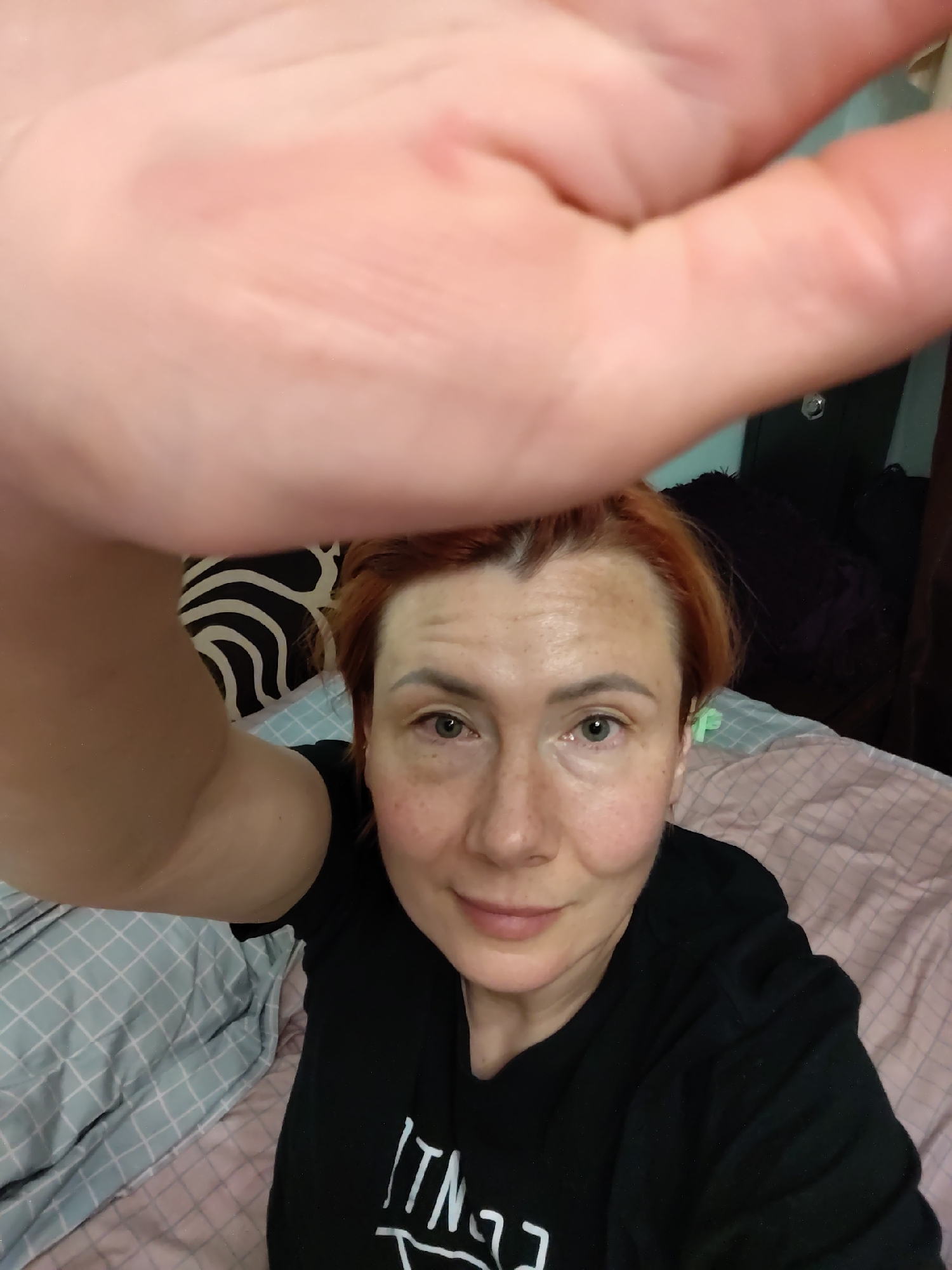 The image is a selfie taken from a close-up perspective, focusing on a woman with reddish-orange hair and fair skin with freckles. She is wearing a black t-shirt. Her right hand is blurred in the foreground, obscuring part of the frame. The background appears to be a bedroom, with a bed covered in pink and light blue patterned bedding. A dark object with a black and white pattern is visible behind the woman's head. The overall scene suggests an indoor setting, likely a home environment. There are no discernible elements that would place the location specifically in Konakovo, Russia.