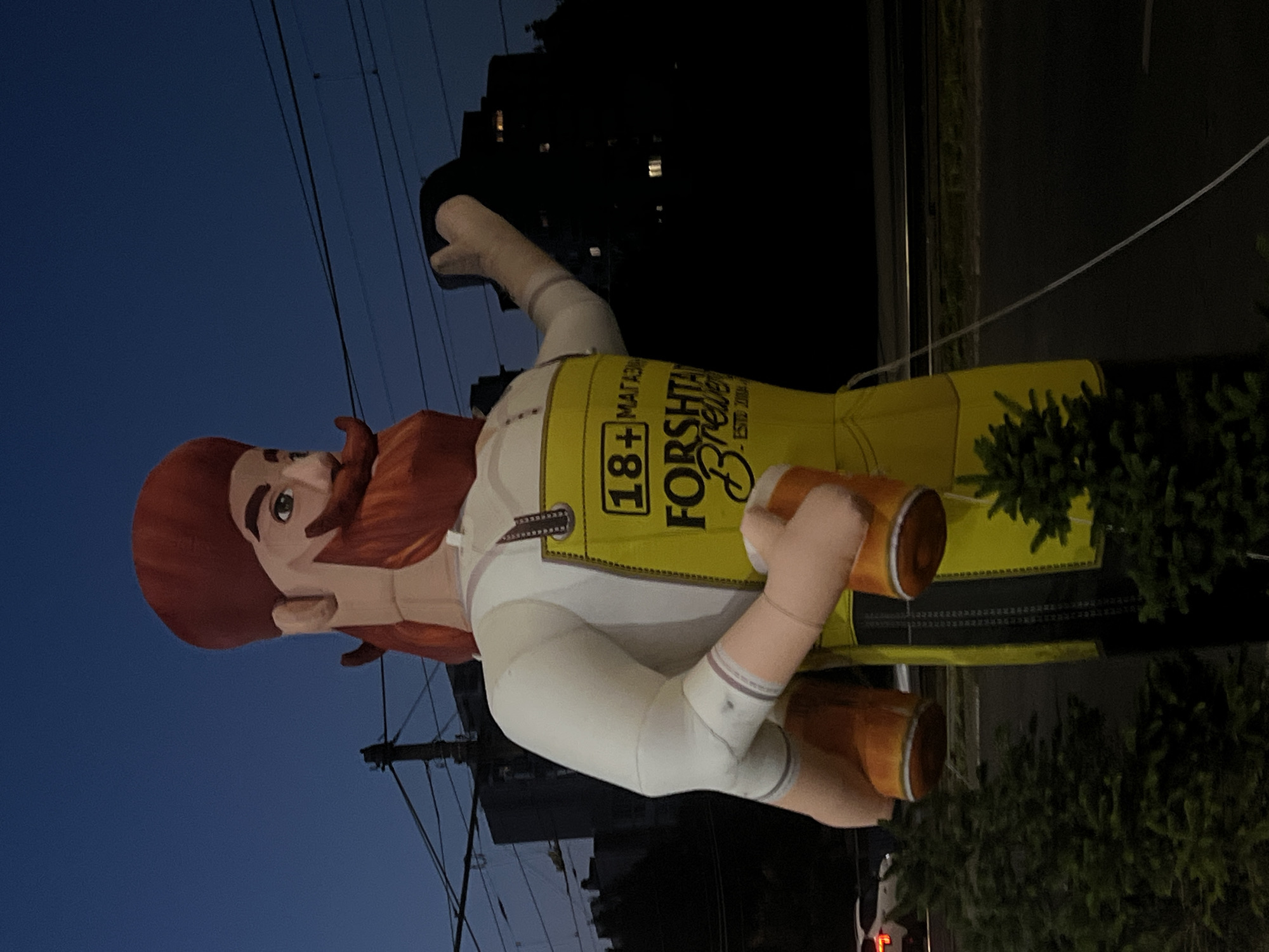 An inflatable figure of a man with a red beard and hat, wearing a yellow apron with "18+ MAGAM" and "Forshtam Brewery - Esto 2004" printed on it, stands outdoors at dusk. The figure is holding two beer mugs. The scene appears to be in Barnaul, Russia, as indicated by the text on the apron. In the background, there are buildings, power lines, trees, and a street with a car.