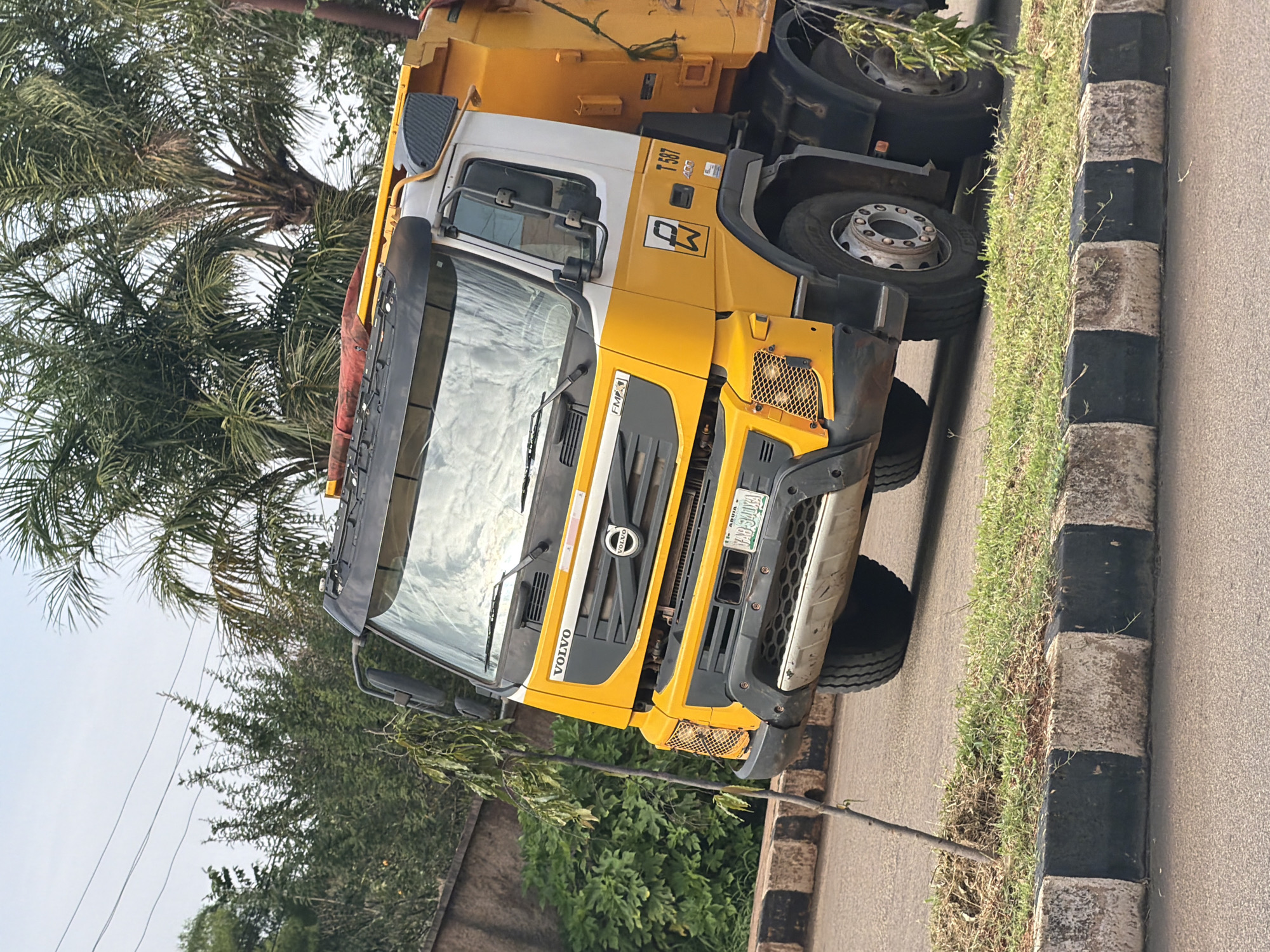 The image shows a bright yellow and white Volvo dump truck parked on an asphalt road in Bukuru, Nigeria. The truck is positioned at an angle, with its front facing towards the left of the frame. A small, thin tree with green leaves leans across the front of the truck. The truck has a prominent "VOLVO" logo on its grille. The license plate is visible and reads "ABUJA YAQ 340 U A". The background consists of lush green trees, including palm trees, and a cloudy sky. A concrete curb with alternating black and white stripes runs along the edge of the road in the foreground. The overall scene appears to be outdoors in an urban or suburban environment.