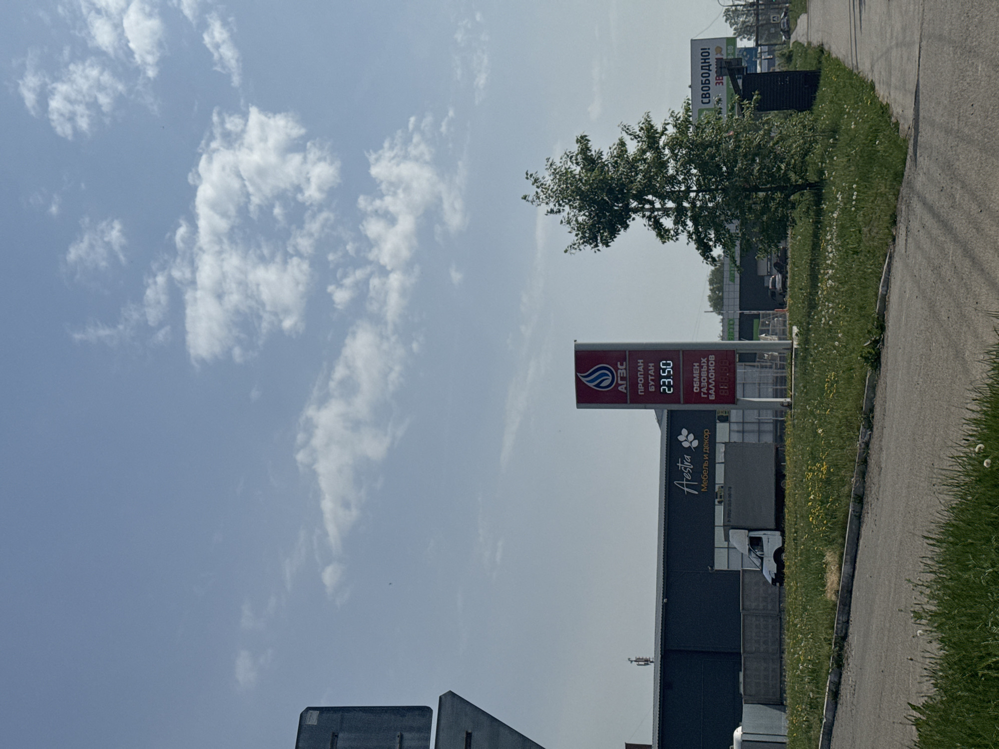 Here is a forensic description of the image:
The image depicts a roadside scene in what appears to be Chaikovsky, Russia. A prominent feature is an АГЗС (Automobile Gas-Filling Station) with a sign indicating the price of propane-butane at 23.50 (likely in rubles). The word "Aestra," possibly a business name in Cyrillic script indicating "Furniture and Decor," is visible on a building. A truck is parked near the building. The sky is mostly clear with scattered clouds. A billboard with Cyrillic text is partially visible in the background. The vegetation consists of grass and a small tree along the edge of the road.
