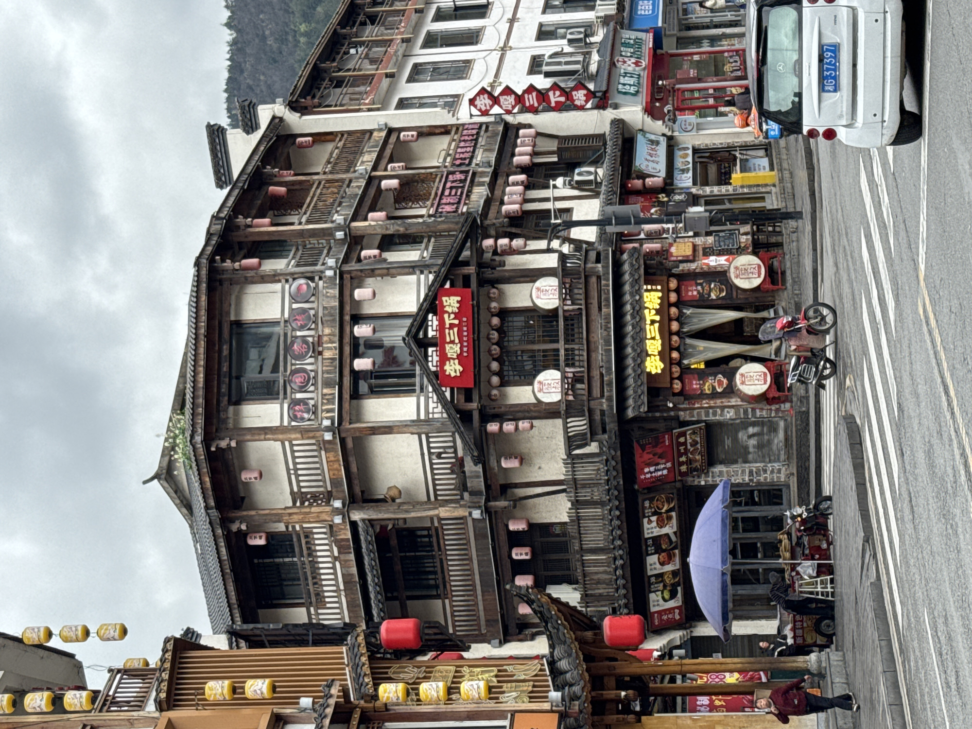 The image depicts a vibrant street corner in Wulingyuan, China, under an overcast sky. A prominent multi-story building, characteristic of traditional Chinese architecture, anchors the scene. It features white stucco walls, dark wooden beams, tiled roofs, and numerous balconies adorned with intricate wooden railings. Red and cylindrical yellow lanterns are suspended extensively from the building's eaves, balconies, and awnings.
The central establishment is a restaurant identified by prominent red signs displaying Chinese characters, most notably "李嘎三下锅" (Li Ga San Xia Guo). The storefronts on the ground floor exhibit various menu boards and decorative elements, including large drums. On the sidewalk in front of this building, a street vendor operates a mobile food stall sheltered by a large blue umbrella. A person in a striped shirt is positioned behind the stall, while another individual in a dark jacket walks past.
The asphalt street features a white striped crosswalk. A white Smart car with license plate 湘G·37397 is positioned on the road, partially obscuring the view of the street ahead. Several motorcycles or scooters, including a red one, are parked near the curb. In the background, forested mountains are visible beneath the grey sky, bordering the urban development. Adjacent buildings continue the architectural theme, featuring a mix of traditional and more contemporary facades with additional commercial signage.