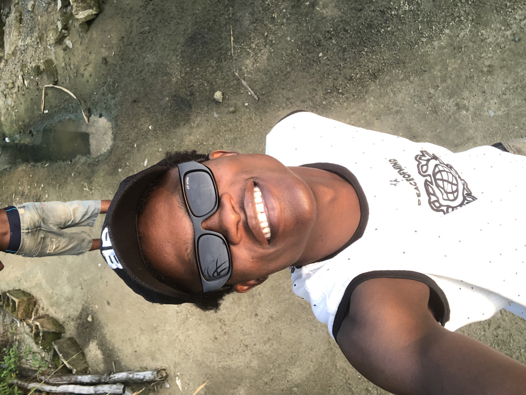 A young Black man in a white sleeveless shirt and a black baseball cap, wearing black sunglasses with "off white" written on the lens, smiles at the camera. He is standing outdoors on a dirt path, with another person in denim shorts and a dark waistband visible in the background. The environment appears to be somewhat unkempt, with scattered rocks, dirt, and a puddle of water. The scene likely takes place in Ado, Nigeria, given the context.