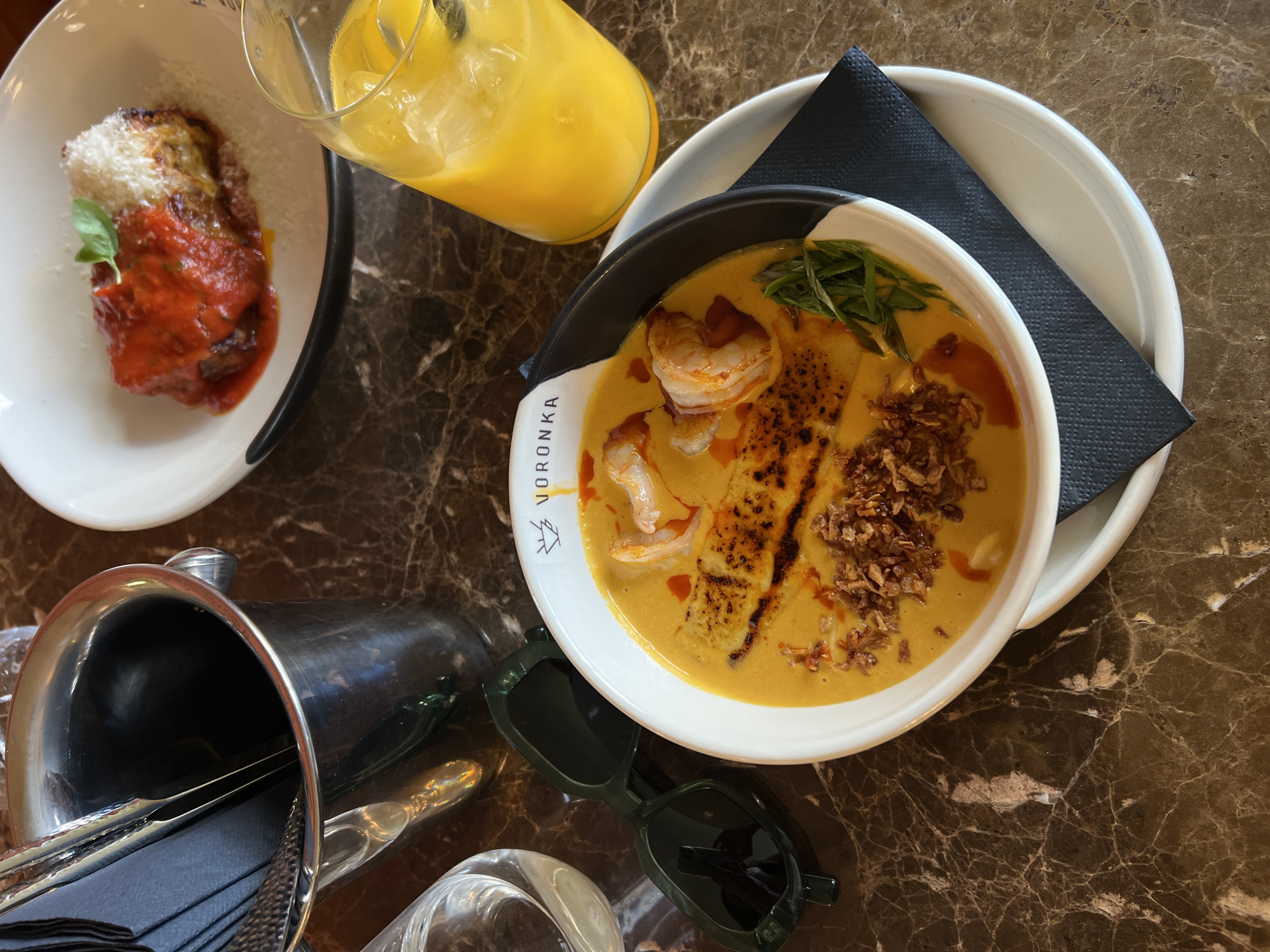 The image depicts a table setting with food and drink. On the left is a white plate containing a dish with red sauce, cheese, and a basil leaf. Next to it is a glass of orange-colored liquid with ice. To the right is a white bowl with a black rim, sitting on a white plate, filled with a yellow soup-like dish containing shrimp, fried shallots, and green garnish, resting on a black napkin. Below, there is a metallic ice bucket containing silverware and black napkins. In front of the bucket is a transparent glass and a pair of dark green sunglasses. The table has a brown marble pattern. The bowl has the word "VORONKA" on it. There is no evidence to confirm it is in the city of Boreti, Montenegro.