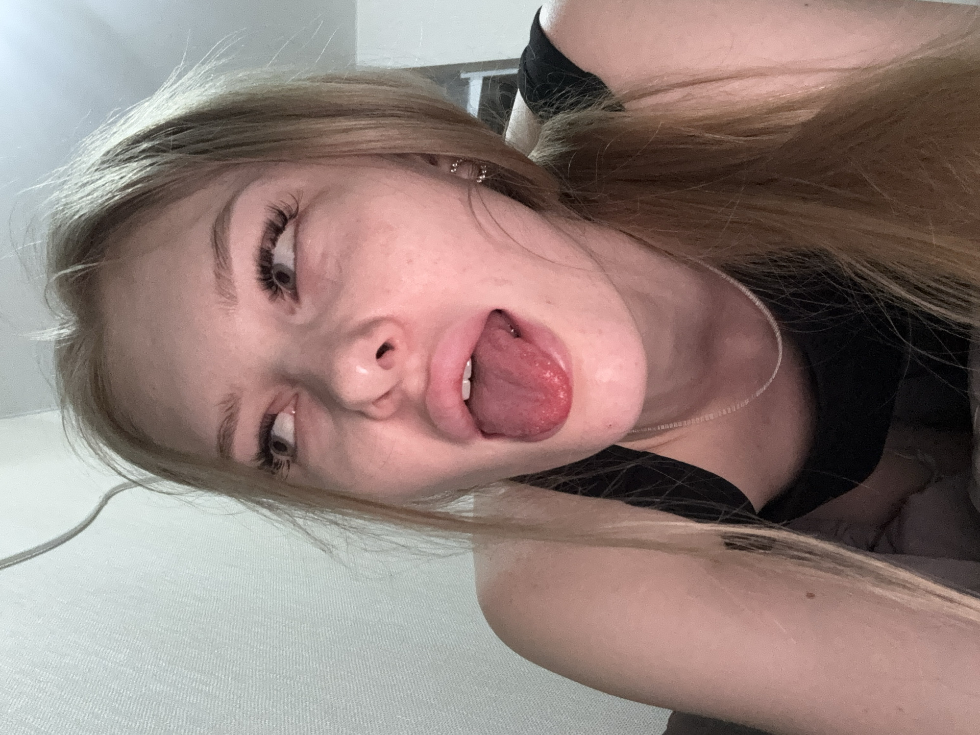 A young woman with long, light brown hair is positioned in a selfie-style photograph. Her head is tilted at an angle, and she is sticking out her tongue. She has light skin, brown eyes, and long eyelashes. She is wearing a black top and a thin silver necklace. A power cable is visible in the lower left corner. There is no definitive information to suggest a location in Perm, Russia. The environment is blurry and indistinct.
