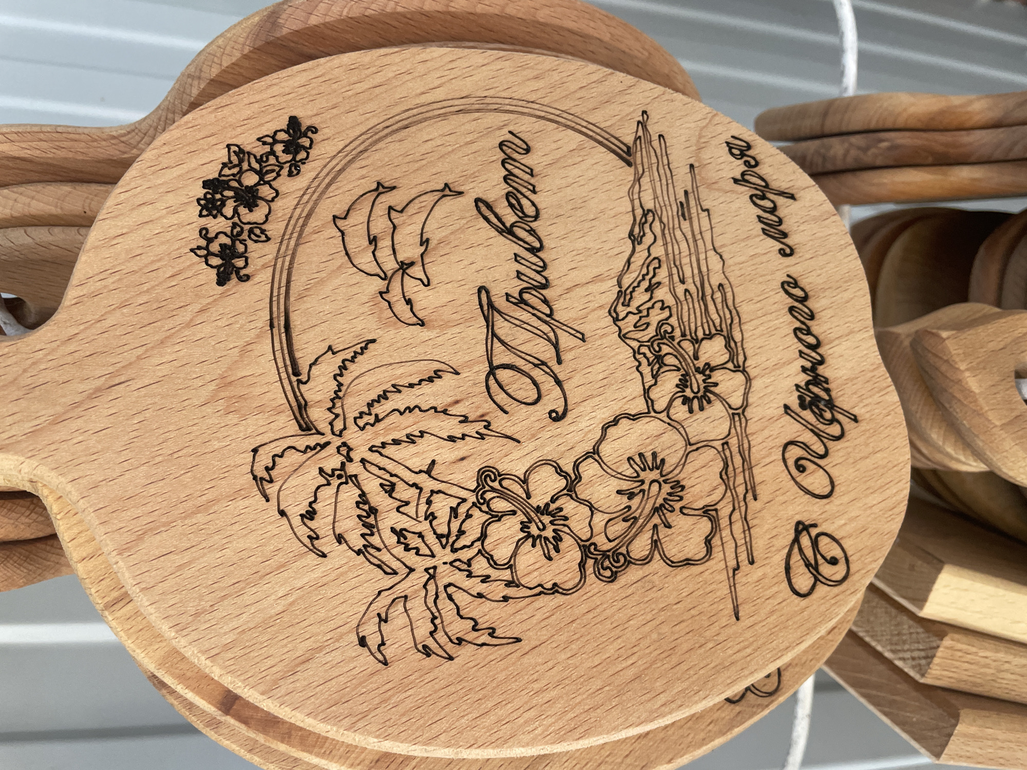 The image shows several round wooden cutting boards stacked together. Each board has a similar design laser-etched onto its surface. The design is enclosed within a circular frame and includes images of dolphins, a palm tree, hibiscus flowers, a seaside cliff, and flowers. There are Cyrillic words etched onto the board in a decorative script. The words are "Привет" and "С Чёрного моря." These translate to "Hello" and "From the Black Sea," respectively. The image was probably taken in Olginka, Russia, based on the translation of "From the Black Sea."