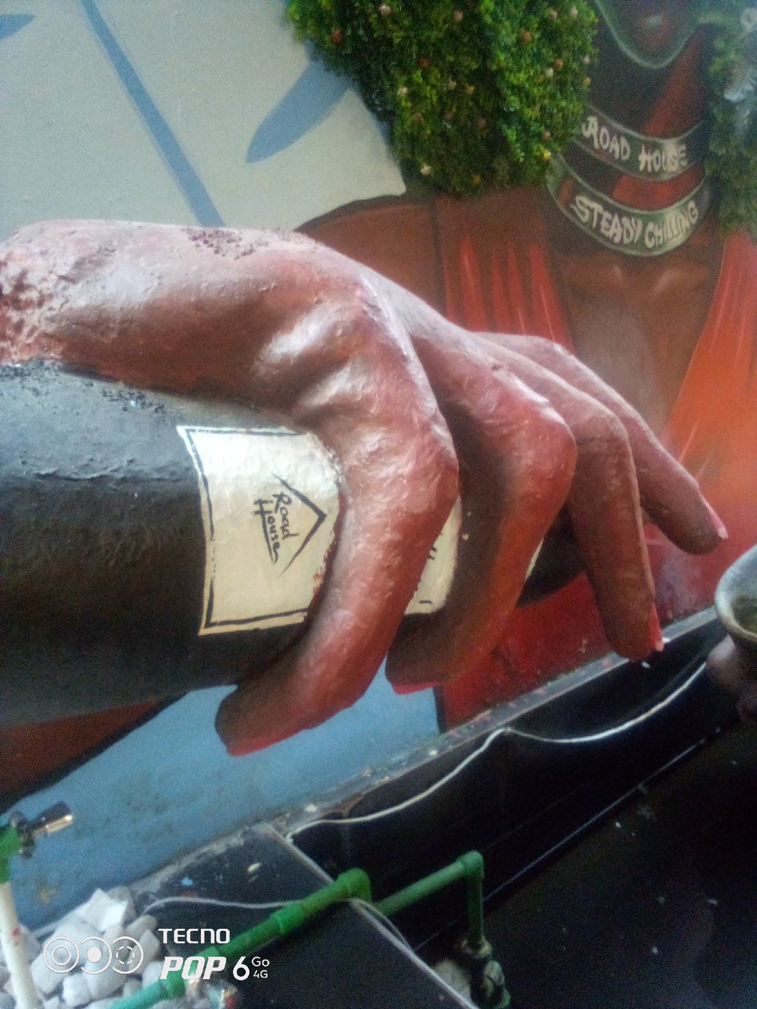 The image depicts a statue of a hand holding a bottle. The bottle has a label with the words "Road House" and "Steady Chilling". The background appears to be a mural or painting, featuring shades of red and blue, with some green decorative elements at the top. The statue of the hand is brown and textured, with visible details of knuckles and fingers. A portion of what appears to be a counter or display surface is visible in the foreground, with some white wires and green pipes. The bottom left corner of the image shows a Tecno Pop 6 4G logo, indicating the device used to capture the photograph. The scene is likely located at a business establishment, possibly a bar or restaurant, given the "Road House" branding. No specific location in Howayi, Nigeria can be definitively identified from the visual information.