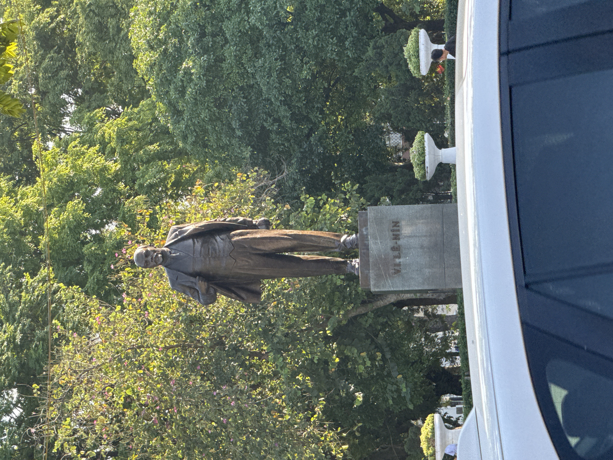 The image shows a bronze statue of Vladimir Lenin in a park setting. The statue is positioned on a gray plinth, and the name "VI LE-NIN" is engraved on the plinth. Lush green trees surround the statue. A white vehicle is partially visible on the right side of the frame. The statue of Lenin is located in Hanoi, Vietnam, near the Vietnam Military History Museum.
