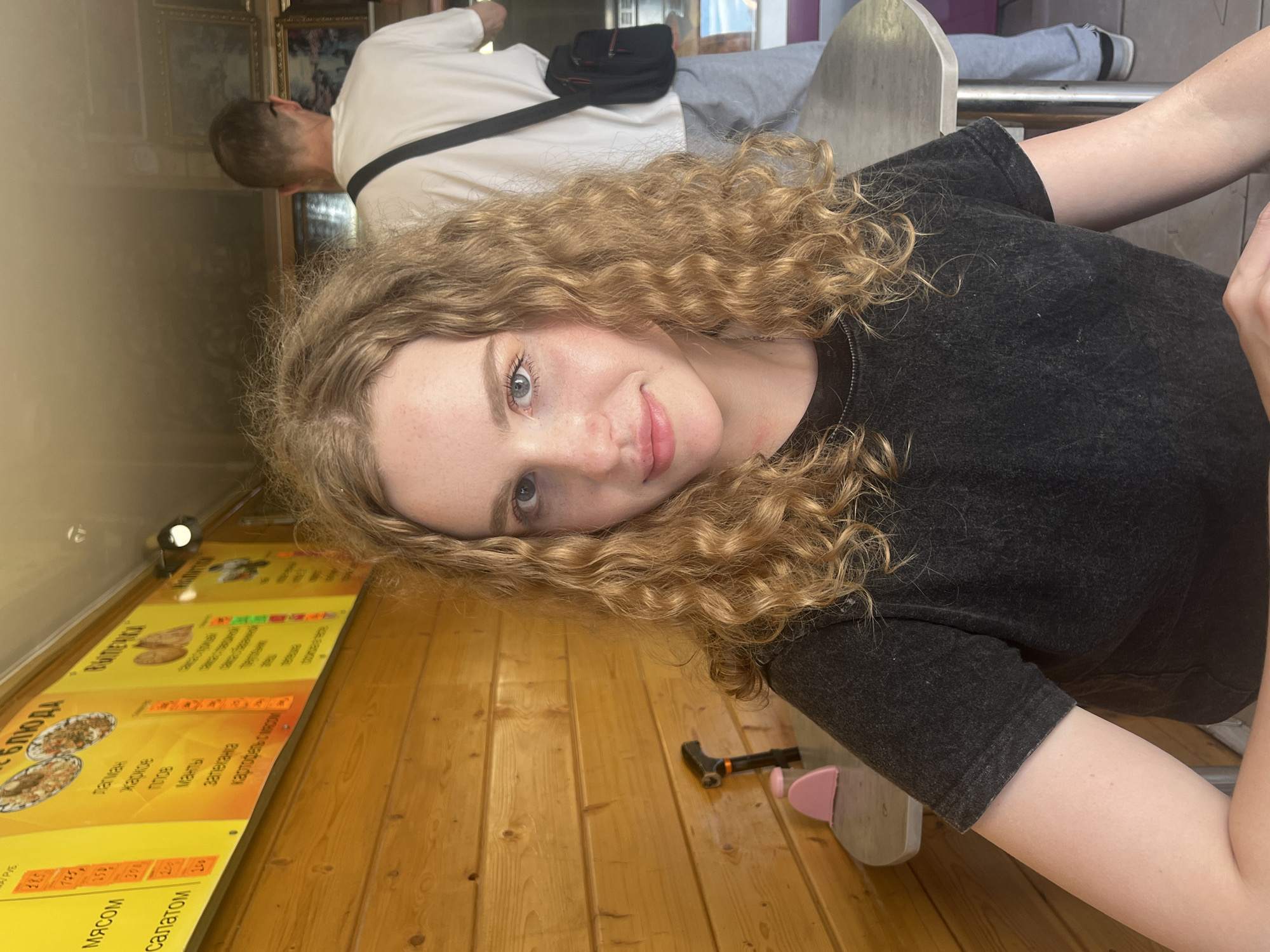 A young Caucasian woman with curly blonde hair and blue eyes is pictured in a selfie. She is wearing a dark grey t-shirt and is sitting at a table. Behind her, a man in a white t-shirt and grey pants is visible, carrying a black bag. The background features a wooden-paneled wall with a menu board displaying Russian text and images of food. To the left of the woman, a black cane-like object and a pink object are on the table. The scene appears to be an outdoor cafe or food stall in Vasilyevo, Russia.