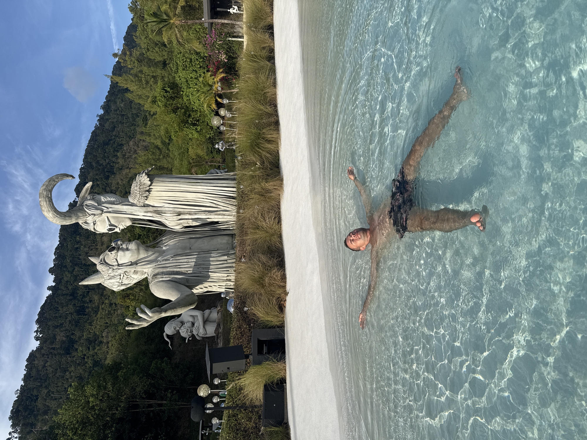 A man floats in a pool in Baan Lipa Noi, Thailand. He is wearing dark shorts and has his arms and legs extended. The pool is located next to a garden with sculptures of figures. A mountain and sky are in the background.