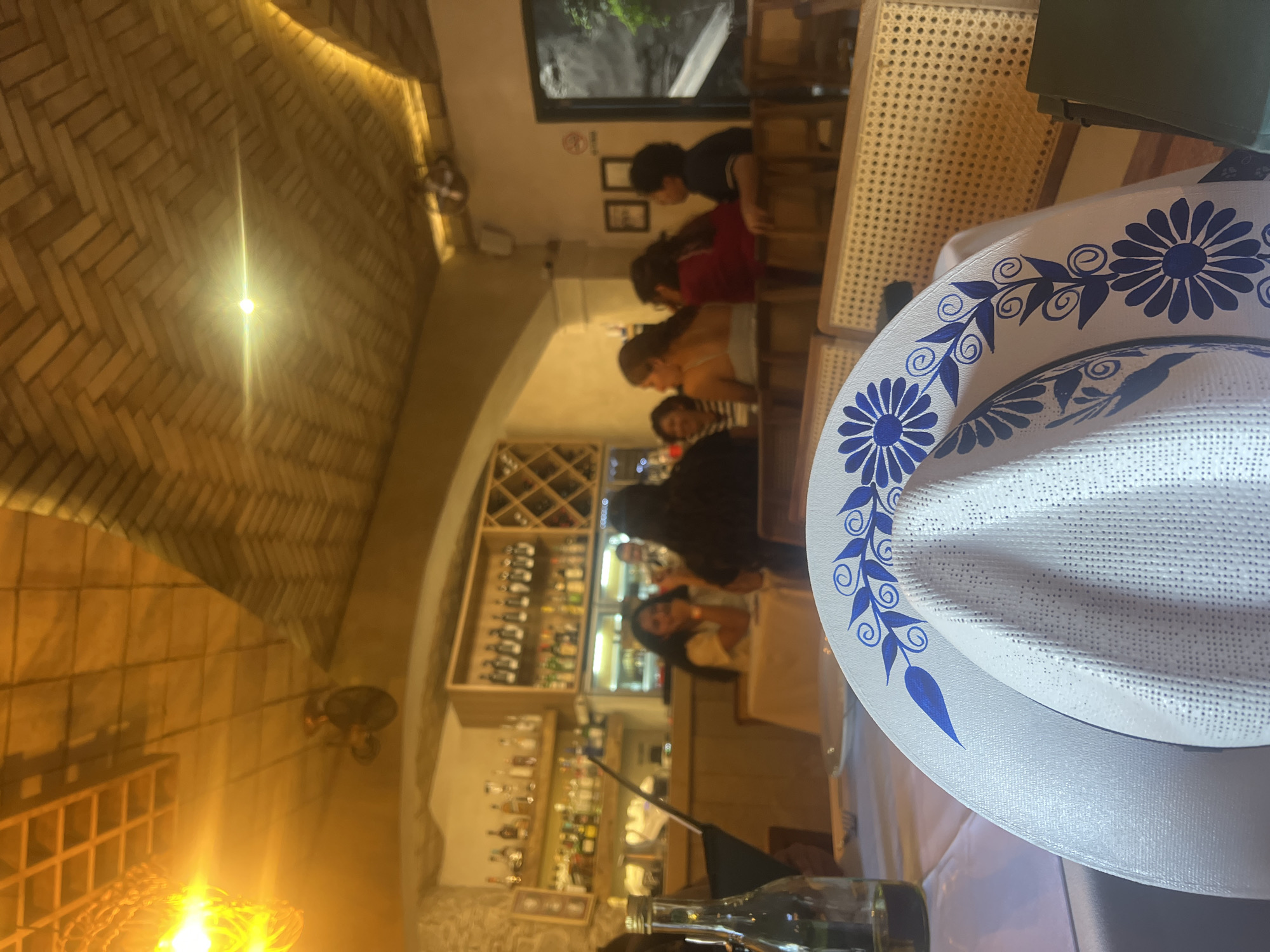 The image depicts the interior of a bar or restaurant in Domingo Arenas, Mexico. The camera focuses on a white sombrero with blue floral patterns on its brim, placed on a table in the foreground. Behind the sombrero, patrons are seated at tables, and some are standing near the bar area. The bar is stocked with various bottles of liquor. The lighting is warm, and the ceiling is finished with a herringbone pattern. The scene suggests a lively social atmosphere.