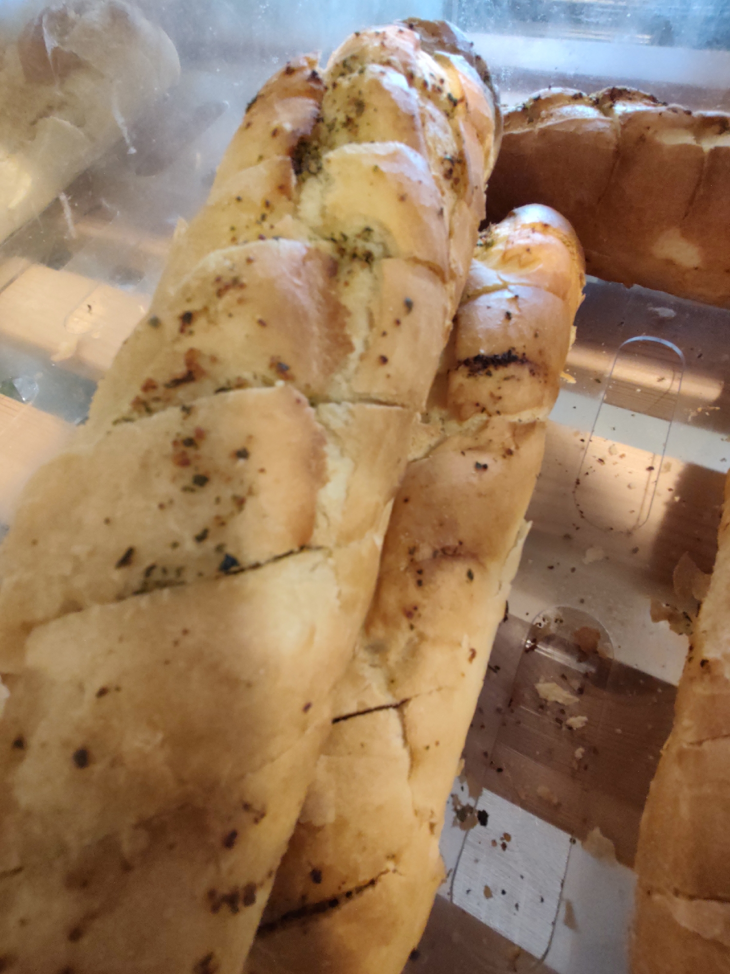 The image shows several pieces of garlic bread in a clear plastic container. The bread is golden brown with visible specks of herbs and spices. The container appears to be on a wooden surface. The background is blurred but suggests an indoor setting, possibly a bakery or a food service establishment. There is no indication of the city of Sochi, Russia, or any people.