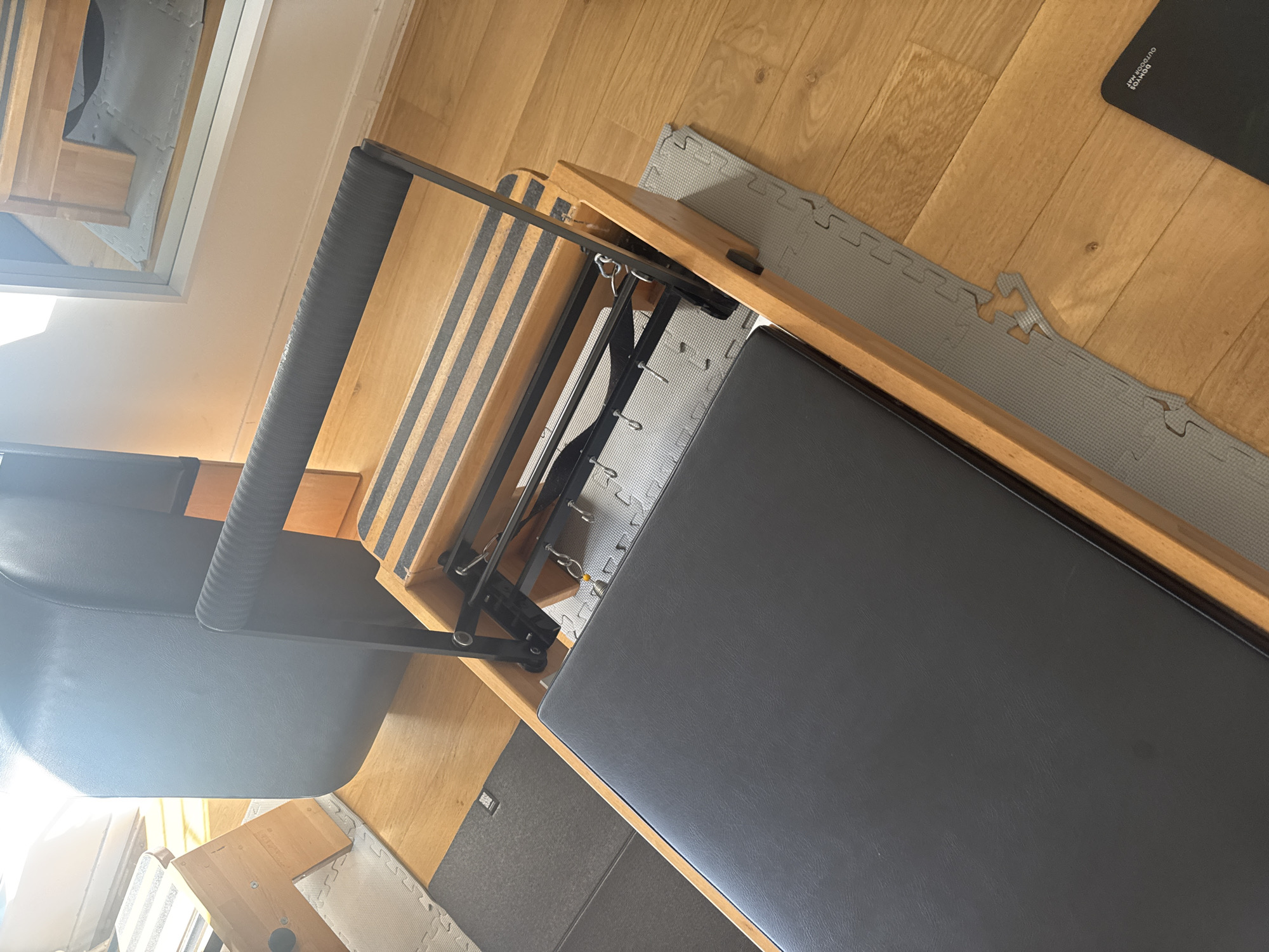 The image shows a pilates reformer machine in a room with light wood flooring and gray foam puzzle mats. The reformer has a wooden frame, a black sliding carriage, and a black headrest. There are black and gray striped springs attached to the carriage. The room has a window in the lower left and a mirror in the upper left. The city of Belgrade, Serbia is not apparent from the visual information provided.