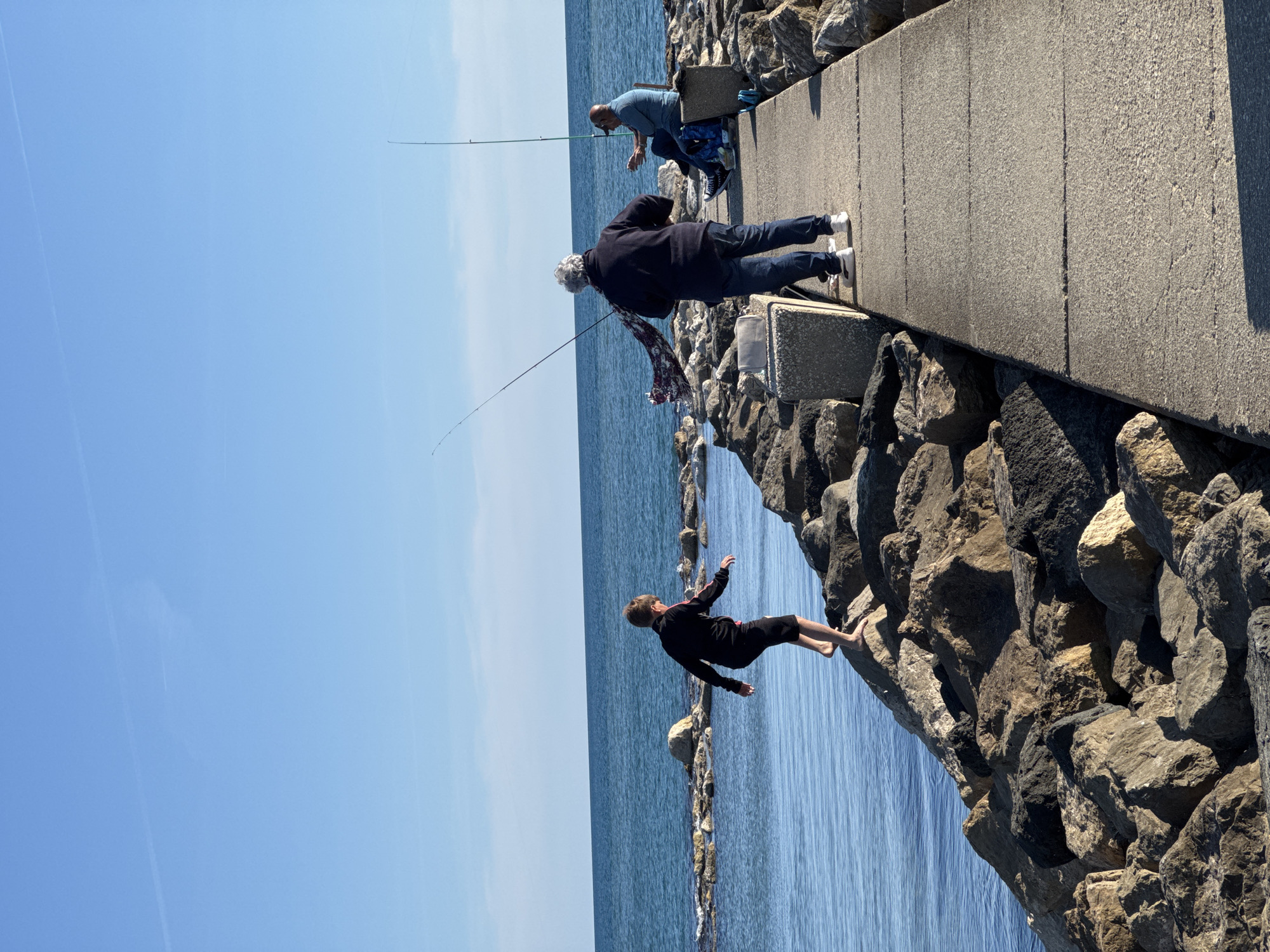 The image depicts a bright, sunny day at a coastal location in Nettuno, Italy, likely a pier or breakwater. The scene features three individuals engaged in leisure activities near the water, set against a backdrop of clear blue sky and calm blue sea.
On a concrete pier or walkway, two men are actively fishing. One man, with grey hair, is standing, dressed in a dark jacket and blue jeans, holding a fishing rod extended towards the water. A patterned cloth or scarf is visible hanging from his side. A second man, with a shaved head and wearing a light blue shirt and blue jeans, is seated on a small concrete block, also with a fishing rod cast into the sea.
To the left of the fishermen, closer to the water's edge and on the rock foundations supporting the pier, a younger individual in a black long-sleeve top and black shorts is positioned. This person is barefoot and appears to be climbing or balancing on the rocks, with their body leaned back and arms slightly outstretched, as if in mid-movement or posing.
The pier itself is a sturdy concrete structure, supported by large, rough-hewn rocks that extend into the water, forming a protective barrier or walkway. The water is clear and appears calm, reflecting the bright sunlight. The sky above is a uniform, vivid blue with no clouds, indicating fair weather. The overall impression is one of a peaceful, sunny day by the sea, enjoyed by people engaged in outdoor recreation. No visible text is present in the image.