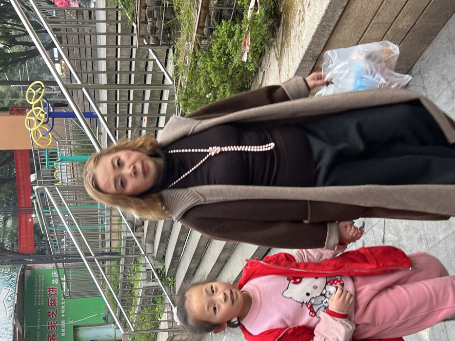 Two individuals, an adult female and a young female child, are depicted walking on a paved pathway outdoors in Jiangjin, China. The adult female, positioned on the right, has light brown, wavy hair and wears a dark brown turtleneck, a long brown and beige overcoat, and dark trousers. A long white pearl necklace, knotted at the chest, is draped over her attire. She holds a transparent plastic bag containing various items in her right hand. Her left hand is joined with the right hand of the child. The child, positioned on the left, has dark hair and wears a pink long-sleeved shirt with a Hello Kitty graphic, pink trousers, and a bright red puffer jacket. Both individuals are looking towards the camera.
The background features a concrete staircase with silver-colored metal railings ascending behind the subjects. To the left of the staircase, a green building with Chinese signage, including "养麦芝麻饼" (Yang Mai Zhi Ma Bing) and "总店" (main store), along with "Jiangjin" in English text, is visible. To the right of the staircase, a small public exercise or play area is partially visible, featuring blue and yellow wheel-like equipment and green structures. A concrete wall with stacked stones and green foliage is present along the right side of the pathway. Red banners are partially visible in the distant background. The scene is illuminated by diffused daylight, suggesting an overcast sky.