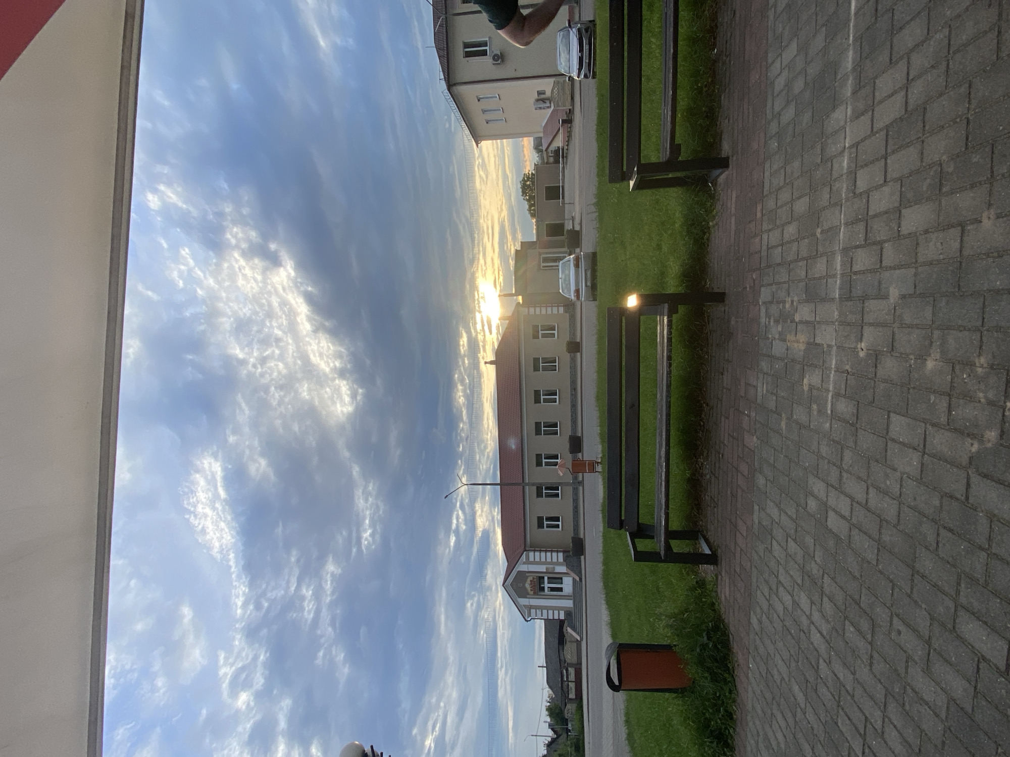 The image captures an outdoor scene with a building in the background and two park benches in the foreground. The building appears to be a single-story structure with a red-tiled roof, large windows, and an entrance with a staircase. Two cars are parked in front of the building. The sky above is filled with dramatic clouds, with the sun setting or rising, casting a warm glow. In the foreground, two wooden park benches are placed on a grassy area, with a paved walkway in front of them. An orange trash bin is visible to the left of the leftmost bench. The overall scene suggests a public space, possibly in a town or city, with an emphasis on an open, communal area. The provided information states the location is Skidzieĺ, Belarus.