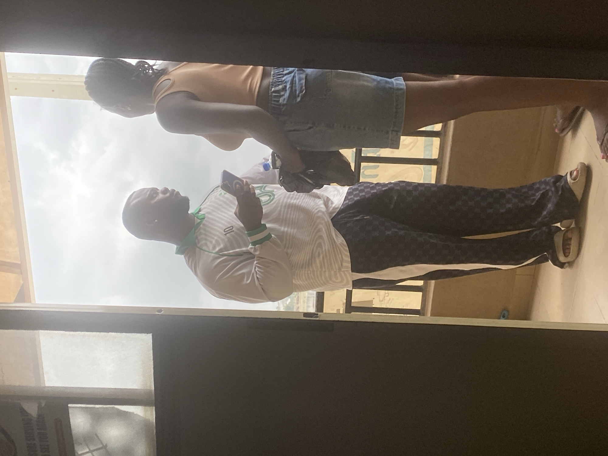 The image, captured in Lugbe, Nigeria, depicts two individuals standing in what appears to be a threshold between an indoor space and an outdoor balcony or open area. The time of day is daytime, under an overcast sky.
On the left, a man is shown from the waist up, wearing a white long-sleeved shirt with green and white striped cuffs and collar, and dark blue pants with a subtle checkerboard or geometric pattern. He holds a smartphone in his right hand and appears to be looking at it.
To his right, a woman with braided hair is visible, wearing a light orange sleeveless top and light blue denim shorts. She stands facing forward, with a small dark bag or object held near her waist.
Both individuals are positioned against a bright, cloudy sky in the background, with a metallic railing or barrier also visible. The immediate foreground shows a dark, possibly wooden or metal, structure at the top and bottom, consistent with a door frame or overhead beam. The floor, partially visible at the bottom right, is tiled.
In the bottom left corner, part of a sign is visible through a screen, with faint, unreadable text that includes fragments such as "SIGNS" and "YOU." The photo is taken from a low angle, looking upwards at the subjects.
