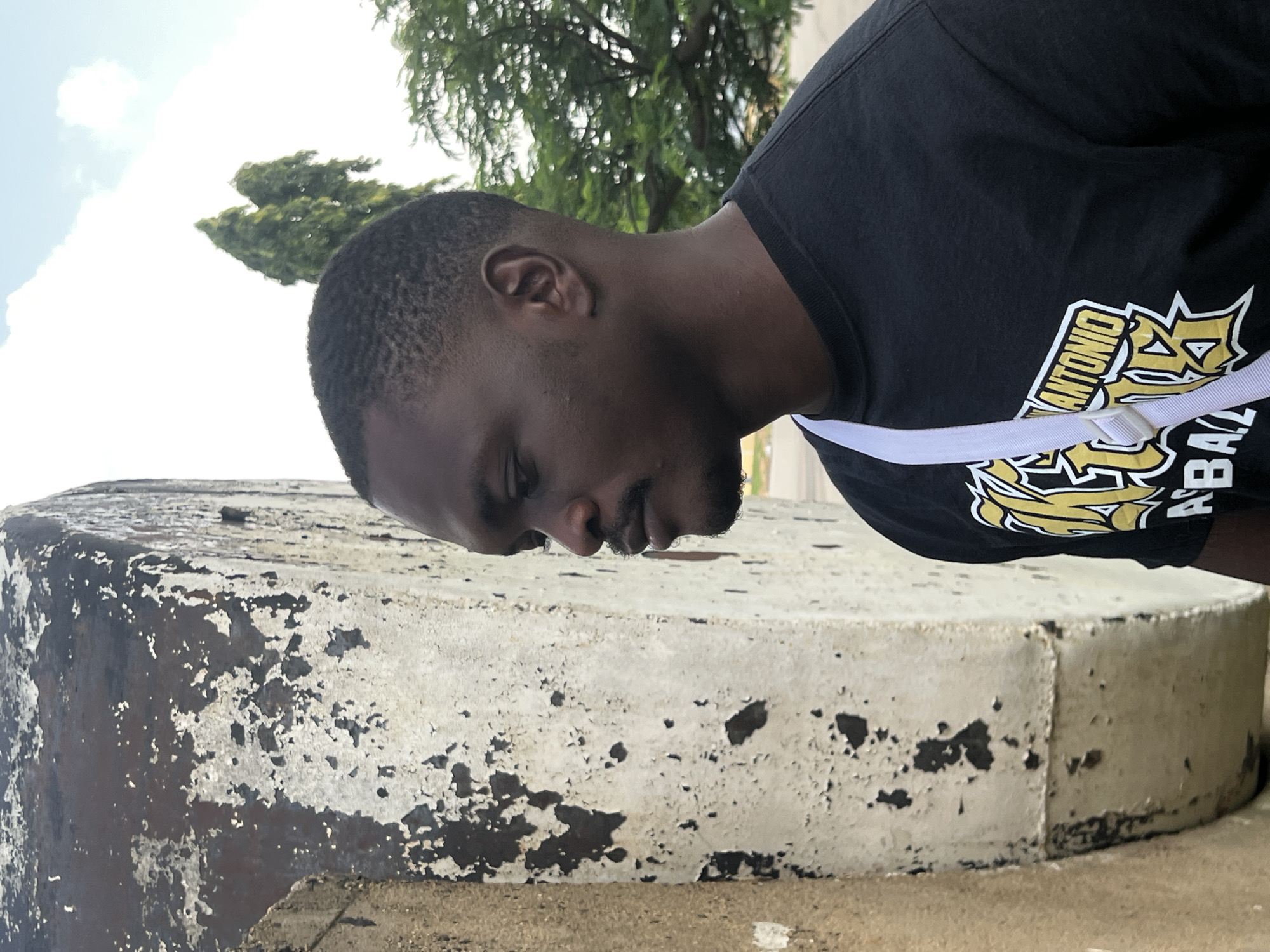 A medium shot shows a young Black man in a black t-shirt with a white strap across his chest. The t-shirt has a yellow and white graphic logo with text that appears to say "SAN ANTONIO" and "BAZ...". He is looking down with his eyes closed and his head is bowed. His right ear is visible, as is the side of his neck and shoulder. He has short hair and a goatee. He is leaning on a large, round, concrete object. The surface of the concrete is worn and has patches of peeling paint, revealing a dark surface underneath. The background shows some greenery, likely trees, and a bright sky with some clouds. The scene appears to be outdoors during the daytime, likely in Jalingo, Nigeria. The man's posture and closed eyes suggest he is resting or in a moment of quiet contemplation.