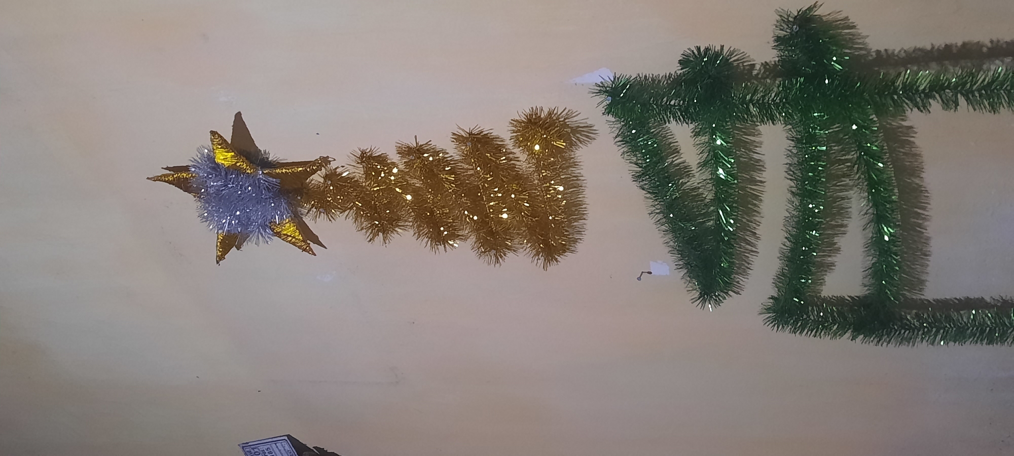 The image shows a wall decoration that resembles a Christmas tree, made of tinsel and lights. At the top, a gold star with silver tinsel is attached. Below the star, a gold tinsel tree shape is formed, adorned with small white lights. Further down, a larger green tinsel tree shape is constructed from draped garlands. The background is a plain, light-colored wall. There are no people visible in the image. The scene appears to be an indoor decoration, potentially in a home or public space. The location in Ploso, Indonesia is not directly evident from the visual information in the image.