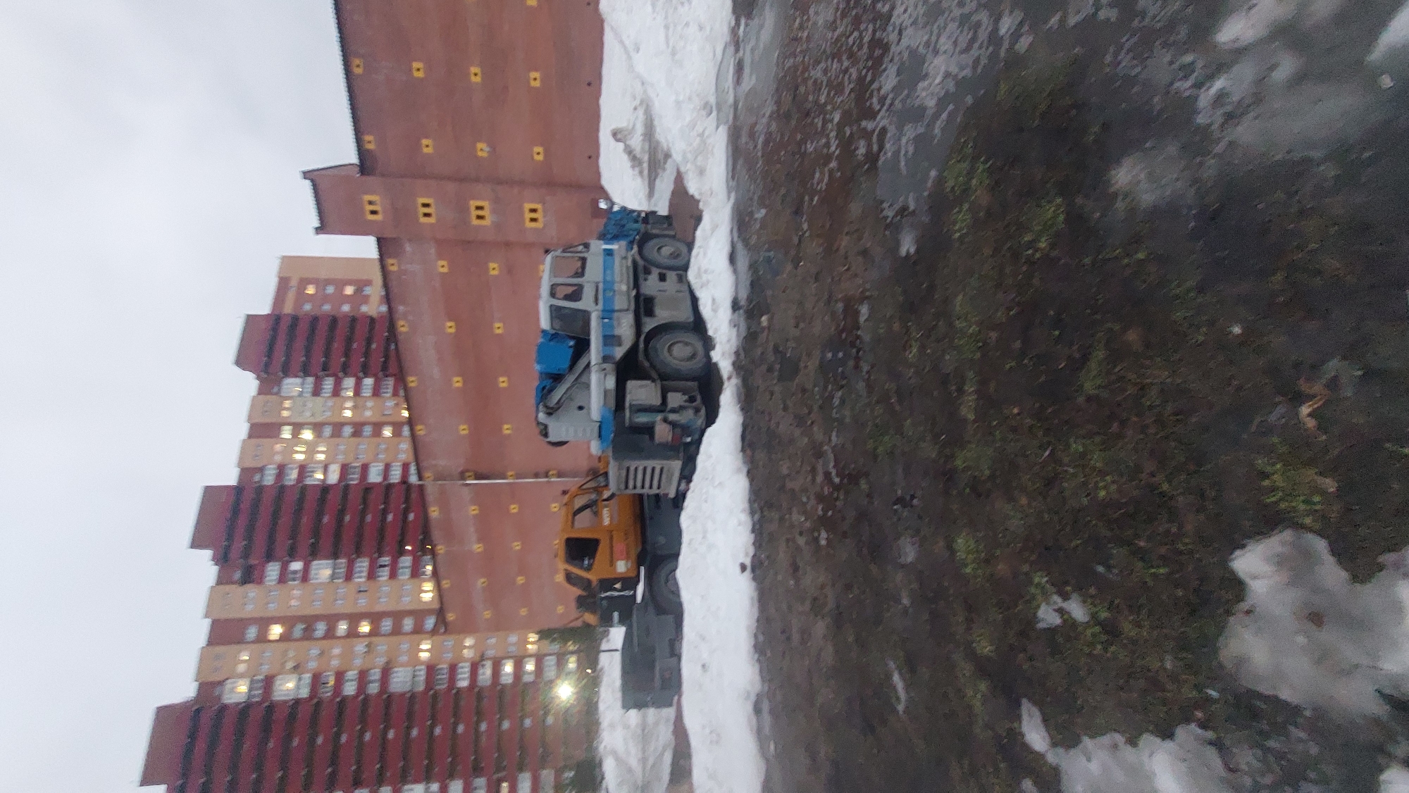 The image depicts a city street scene in Novosibirsk, Russia. Two construction vehicles, possibly cranes, are parked adjacent to a large building complex. Patches of snow and wet ground indicate a recent or ongoing thaw. The buildings are tall with a multi-tiered, patterned facade, alternating between darker and lighter colored sections. The sky is overcast, suggesting a grey day. No people are visible in the frame.