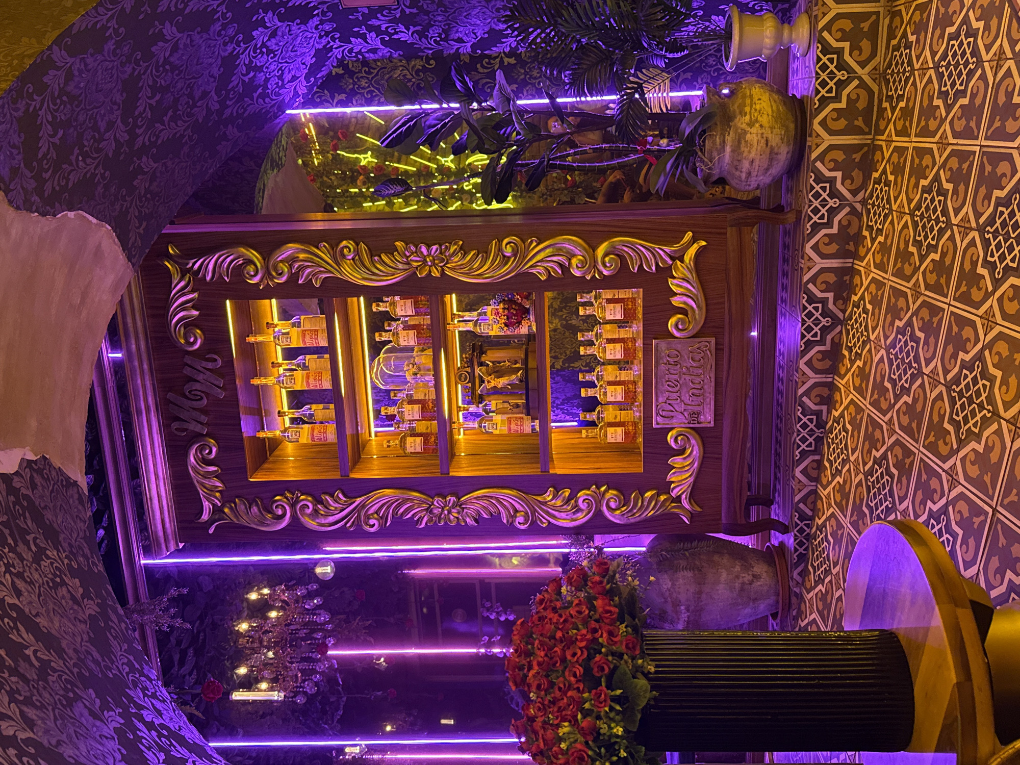 A display shelf with ornate gold trim is illuminated by neon purple lighting. The shelf contains bottles, and the words "Puerto Indias" are visible. The surrounding decor includes patterned wallpaper and tile work. A plant and a ceramic vase are positioned above the shelf. A large vase filled with red roses is in the foreground. The location is possibly Puebla, Mexico based on the image hints.