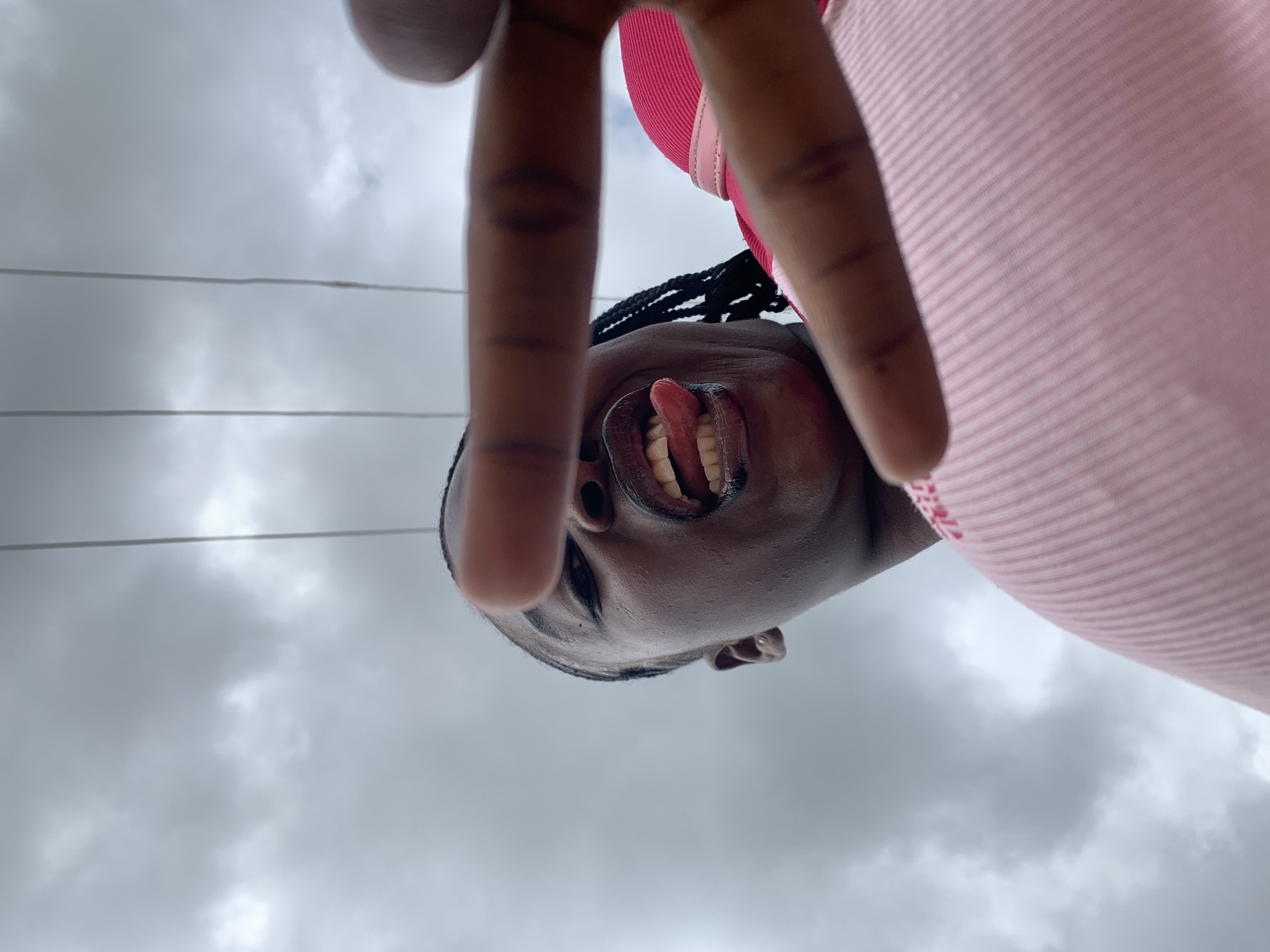 An upward-angle, close-up, self-portrait captures a young Black woman from the chest up against a cloudy sky. She has dark skin, dark braided hair, and is wearing a pink garment that appears to be a top or dress. Her mouth is open, and her tongue is sticking out. Her right hand is extended towards the camera, obscuring part of her face, with her index finger and thumb forming a peace sign gesture. Two power lines are visible in the sky above her. The image does not contain specific information to identify a location in Jos, Nigeria.
