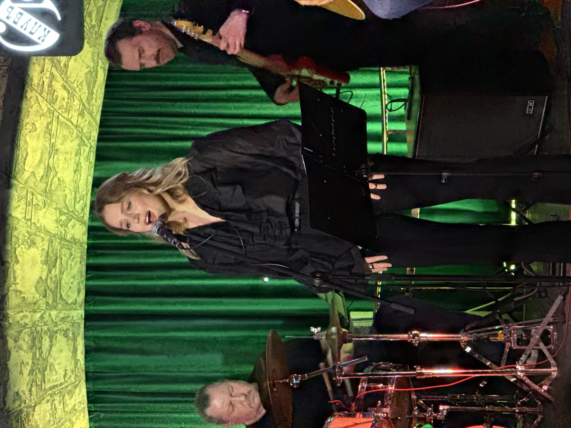 This image captures a live musical performance by a band in an indoor venue, likely a club or bar in Moscow, Russia. This location is suggested by the Cyrillic text "КЛУБ" (KLUB), meaning "club," visible on a backlit sign on the left side of the frame. The artificial lighting indicates that the event is taking place in the evening or at night.
Three musicians are central to the scene. In the foreground, slightly to the right, a female vocalist with blonde hair, dressed in black, sings into a microphone. Her mouth is open, suggesting she is mid-song, and a music stand with sheet music or lyrics is positioned in front of her. Behind her and to the right, a male guitarist, also in black, plays an electric guitar, focused intently on his instrument. In the lower left corner, a male drummer is seated behind a drum kit, with cymbals and drums visible as he performs.
The stage is set against a backdrop of dark green, horizontally draped curtains. To the far left, a textured, light-colored wall, possibly stone or brick, is visible, adding architectural detail to the venue. An amplifier can be seen on the stage floor to the right. The overall impression is an engaging and focused live music performance in a casual club setting.
