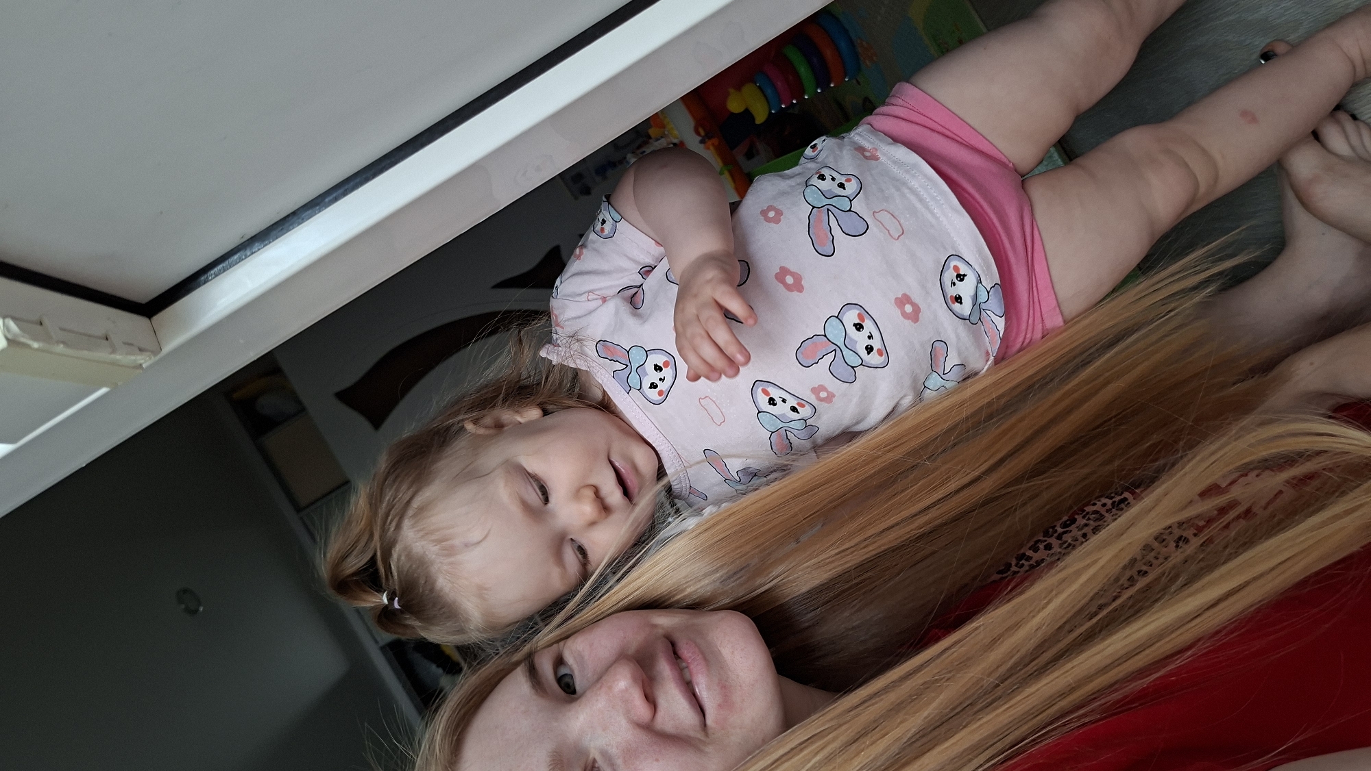 The image depicts a selfie taken indoors in what appears to be a private residence. A light-skinned woman with long, blonde hair, wearing a red shirt, is looking at the camera. Beside her, a young child, presumably a baby, lies on their back. The baby is wearing a light pink outfit with a pattern of white bunnies and flowers, and pink shorts.
Part of a white piece of furniture, possibly a cabinet or shelf, is visible in the upper left corner of the frame. In the background, there's a colorful play mat with various toys. The floor seems to be of a gray, wood-like material.
While the image does not inherently suggest any connection to Chaikovsky, Russia, the presence of that location in the prompt suggests it might be relevant context known outside the image. Without that prior information, the image itself provides no clues about the location.