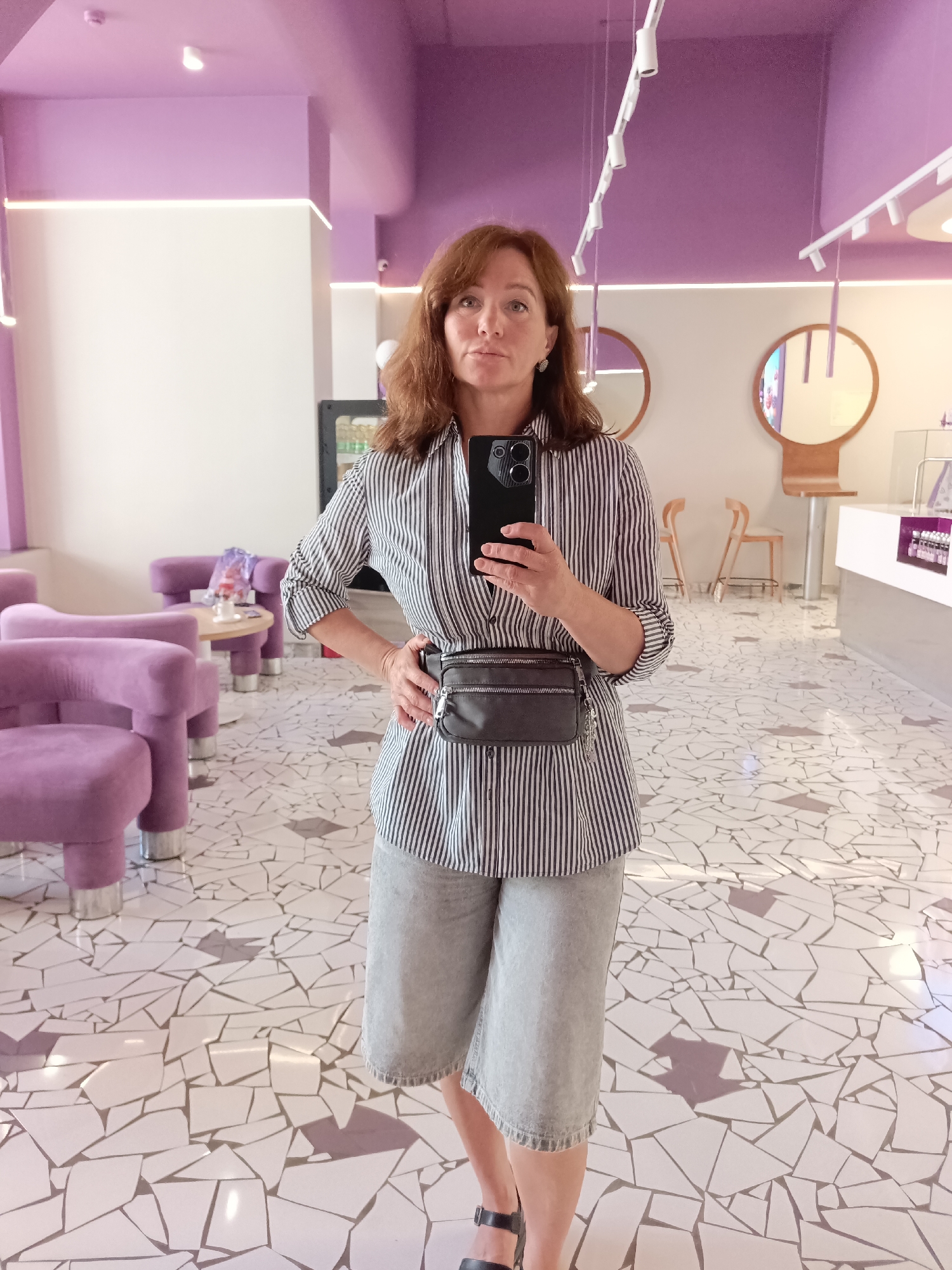 A woman stands in a brightly lit, modern establishment with a pink-purple ceiling and white walls, taking a selfie in a mirror. She is wearing a black and white striped button-down shirt, grey denim shorts, and black sandals. A dark gray fanny pack is worn across her waist. Her left hand is on her hip, and her right hand holds a smartphone. The floor is covered in a mosaic of irregularly shaped white and grey tiles. In the background, to the left, are several plush purple armchairs around a small round table. To the right, a counter area is visible with shelves holding products. The overall aesthetic suggests a cafe or boutique. The image was taken in Saratov, Russia.