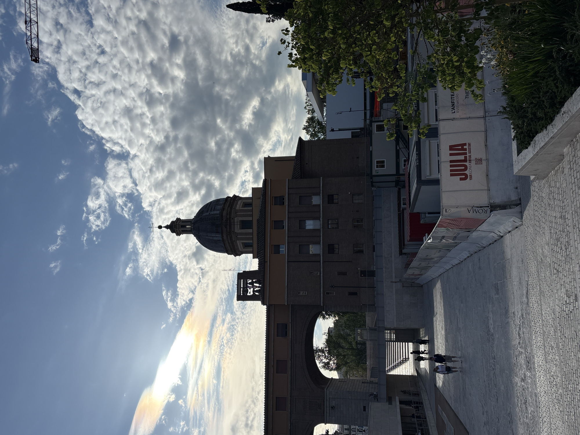 This image captures an urban scene in Rome, Italy, under a partly cloudy, bright sky. A large, grey architectural dome, possibly belonging to a church or historic building, dominates the left side of the frame, topped with a cross-like finial. Below it, a multi-story brick building features a bell-like structure and a grand, wide archway that provides a glimpse of trees and buildings beyond.
On the right, a series of stacked, white container-like structures are visible. These units display prominent red text reading "JULIA" and "YOUR ROMAN VIRTUAL ASSISTANT," along with a QR code. The word "ROMA" is also visible at the base of these containers. Further text on these structures, "L'ANFITEA..." and "THE XVIII SECOLO," suggests proximity to an 18th-century amphitheater. A large construction crane boom is partially visible against the sky in the upper left.
In the foreground, a wide, paved area stretches across the bottom, with a textured stone wall along the right edge. Several people are seen walking away from the viewer on this paved surface, appearing as dark silhouettes.
The time of day is likely midday or early afternoon, characterized by intense sunlight. This light creates a noticeable lens flare and a vibrant circumhorizontal arc (or sun dog) effect in the clouds in the upper left sky. The scene blends historical Roman architecture with contemporary urban elements and activity.