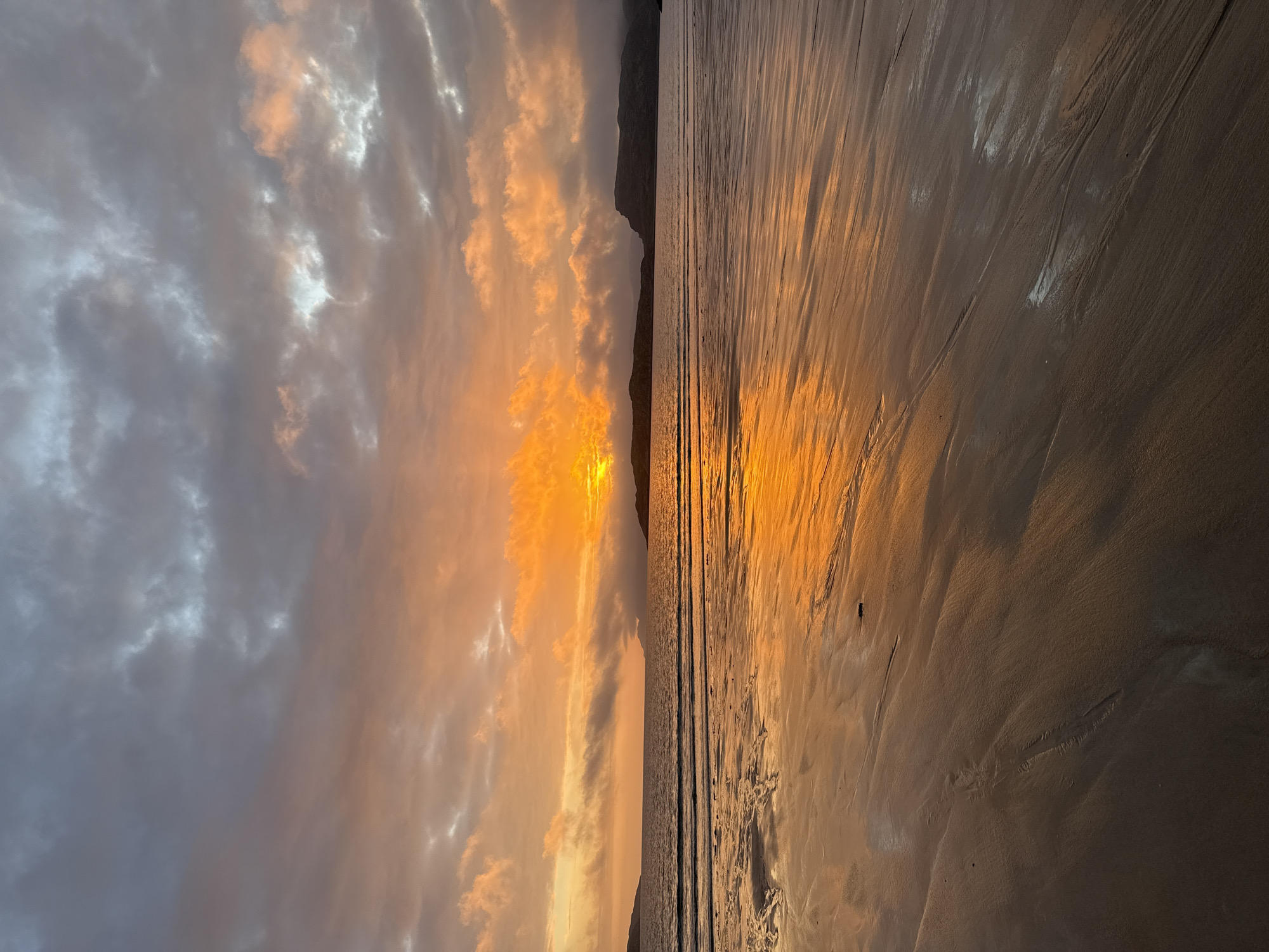 A dramatic sunset paints the sky in hues of orange and grey over a calm sea and a wet sandy beach in Teriberka, Russia. The sun, partially obscured by clouds, casts a vibrant orange glow that reflects on the wet sand, creating a mirror-like surface with intricate patterns of water and sand. Dark, hilly landforms are visible in the distance on both sides of the frame, silhouetted against the luminous sky. The clouds are a mix of dark grey and bright, illuminated by the setting sun. The scene is devoid of any people or animals, presenting a serene and isolated natural landscape.