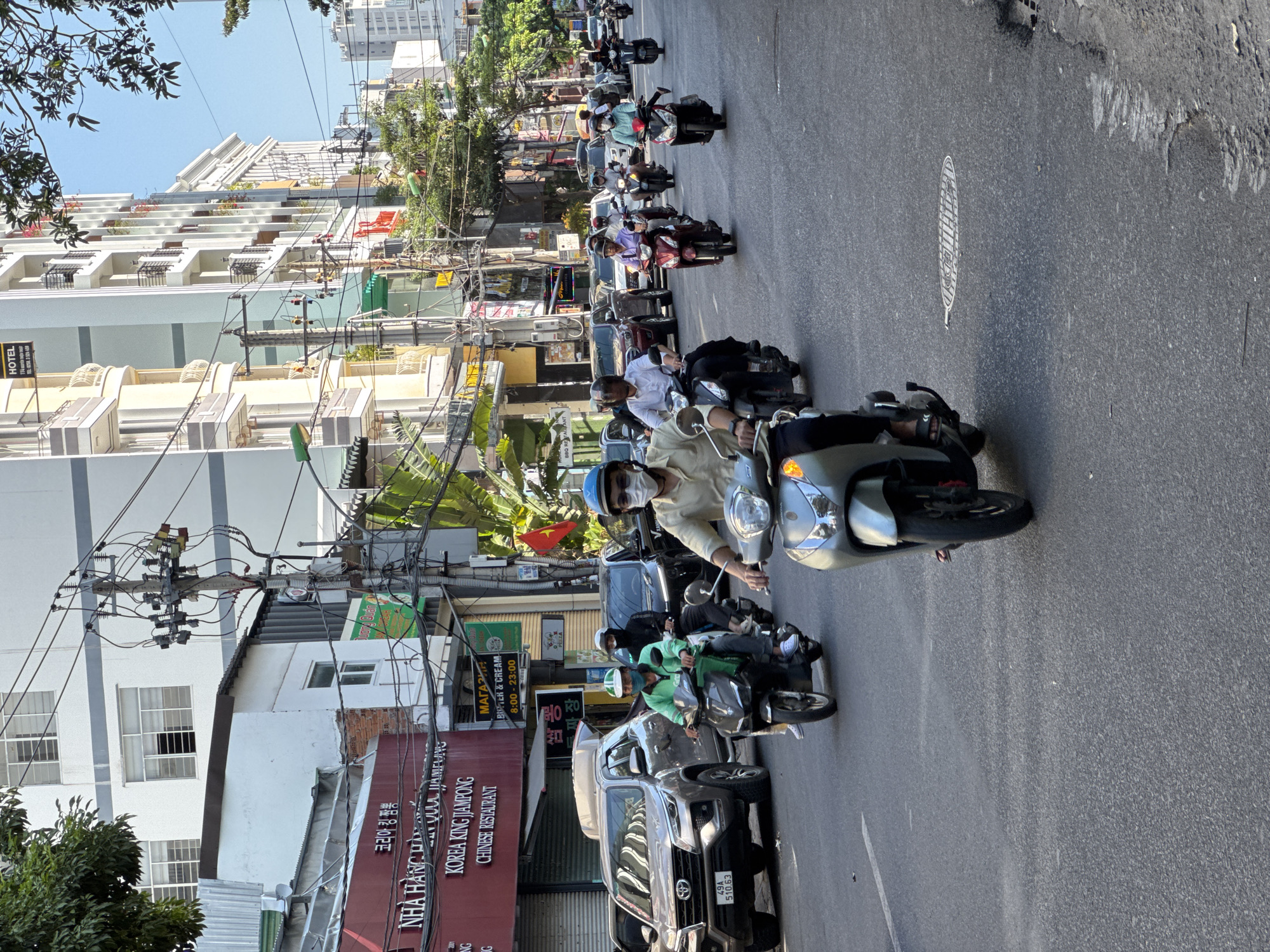 This is a street-level, medium-distance, eye-level shot of a busy street in Nha Trang, Vietnam. The image is dominated by a traffic scene with multiple motorbikes and a few cars. The foreground features a person riding a silver scooter, seen from the chest up, wearing a light-colored shirt, a light blue helmet, and a white face mask. Behind them, another scooter rider in a white helmet and dark shirt is visible.
In the mid-ground, several other scooter riders are navigating the road. One rider in a green shirt and white helmet is on a black scooter, and another in a white shirt and helmet is on a dark scooter. To the left, a silver SUV is parked. Further down the street, more vehicles and buildings are visible, including a multi-story building with balconies and a large number of electrical wires and poles lining the street. Signs with Vietnamese and Korean text are visible on the buildings, suggesting a commercial area.
The background shows more buildings, trees, and a clear blue sky, indicating daytime. The overall impression is one of bustling urban traffic in a Southeast Asian city.