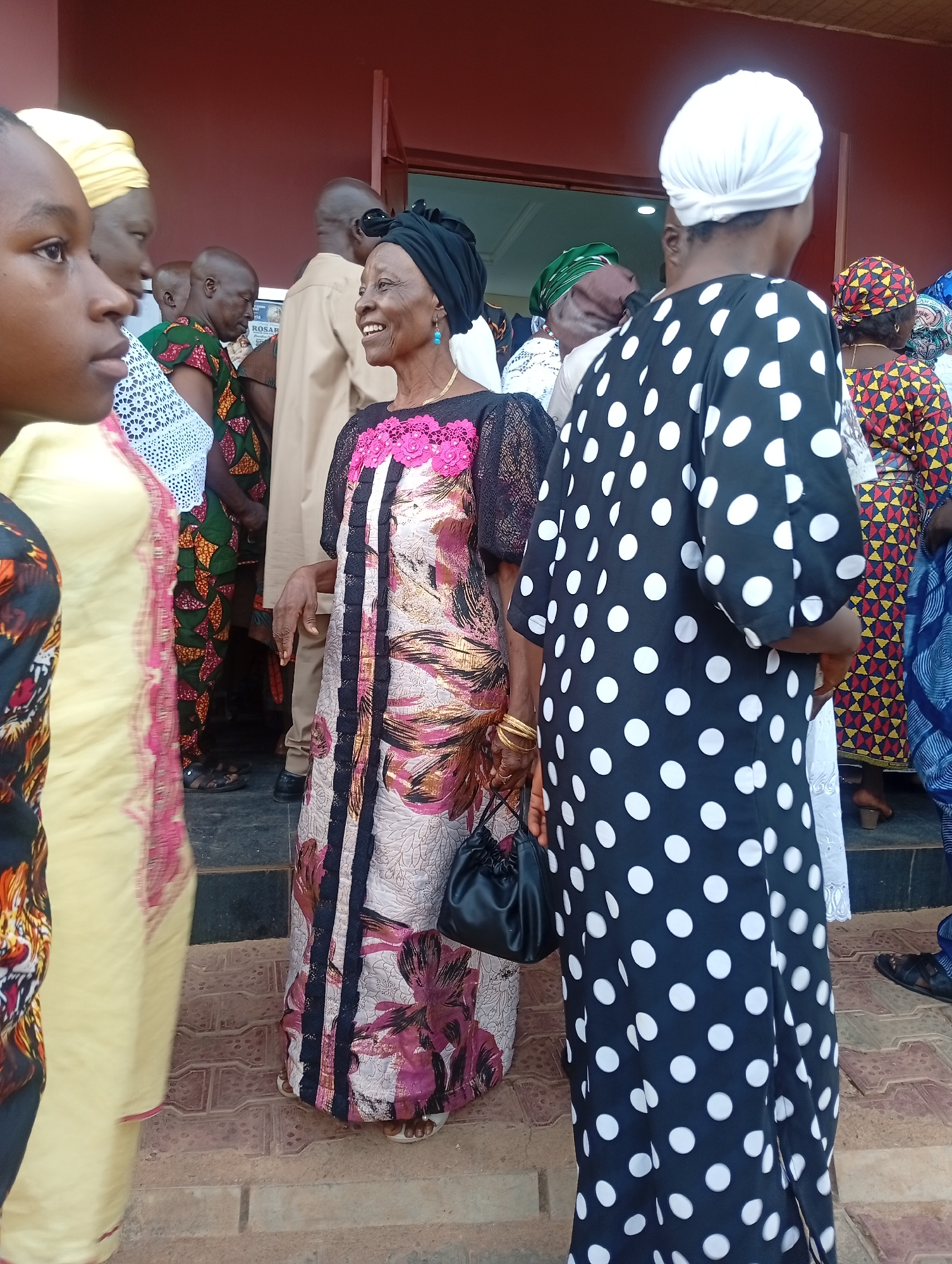 A group of people stand near an entrance, likely in Narayi, Nigeria. A woman in the center smiles, wearing a patterned dress, black headwrap, gold bracelets, and carrying a black purse. To her right, a woman wears a black and white polka-dot dress and a white headwrap. Others in the group wear various patterned clothes. The setting appears to be a building with a red exterior.