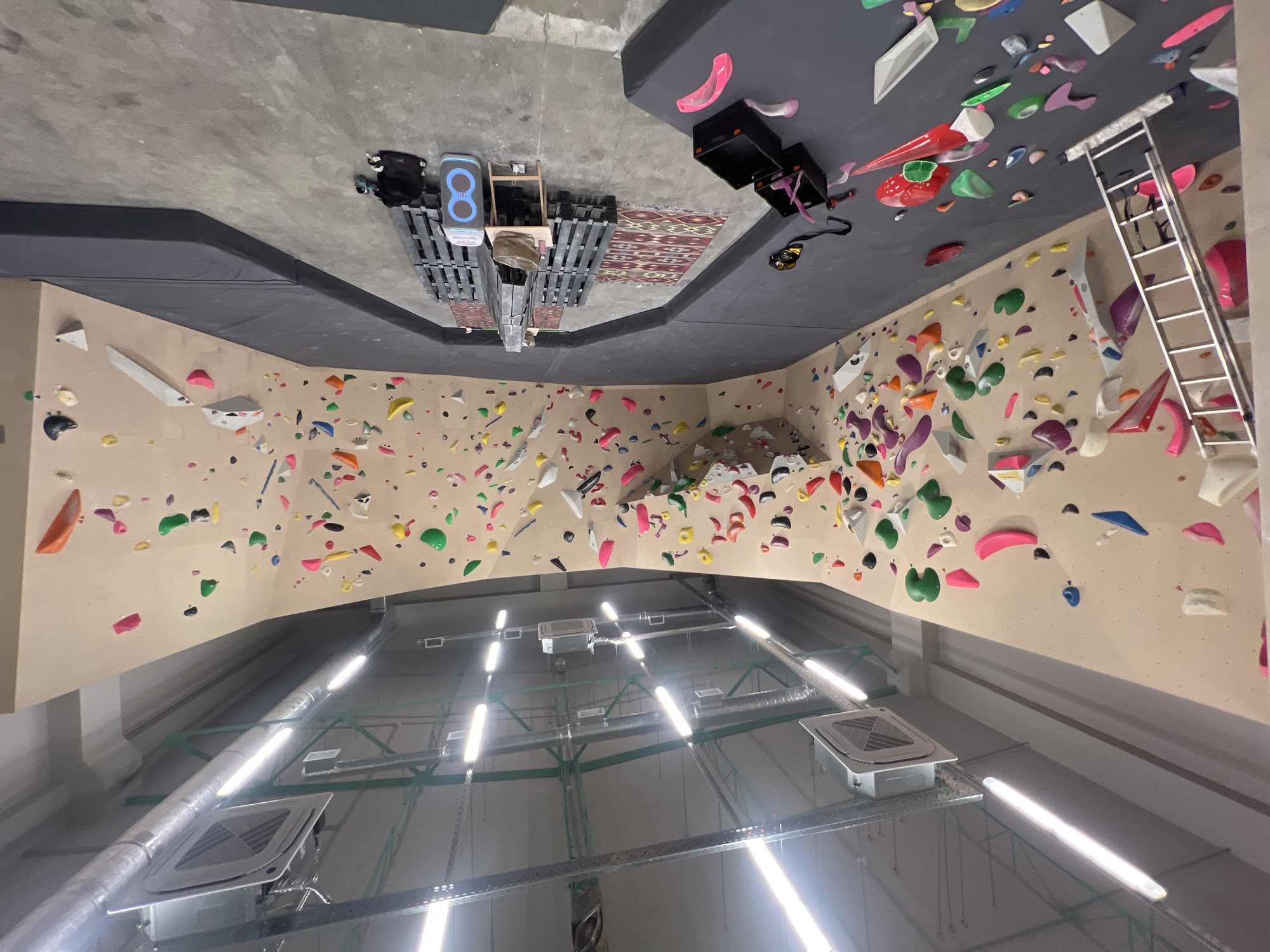 The image shows the interior of a climbing gym in Sochi, Russia. The walls are covered with colorful climbing holds. A safety mat covers the area below the climbing walls. A ladder leaning against one wall provides access to higher holds. The ceiling is high, with fluorescent lights and ventilation systems visible. Miscellaneous objects are situated on the floor in the distance. The orientation of the image is rotated.