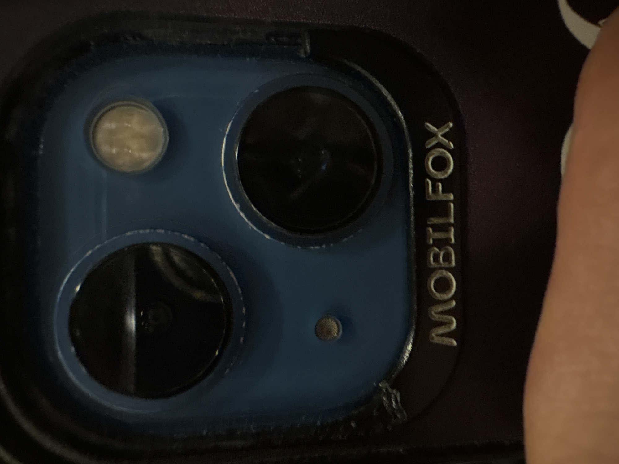 The image shows a close-up of a cell phone camera module with a Mobilfox branded protective case. The camera module consists of two larger lenses, a smaller lens for the flash, and a small microphone. The case is dark-colored with gold lettering. Part of a finger is visible on the right side of the image. The location is not discernible from the image.
