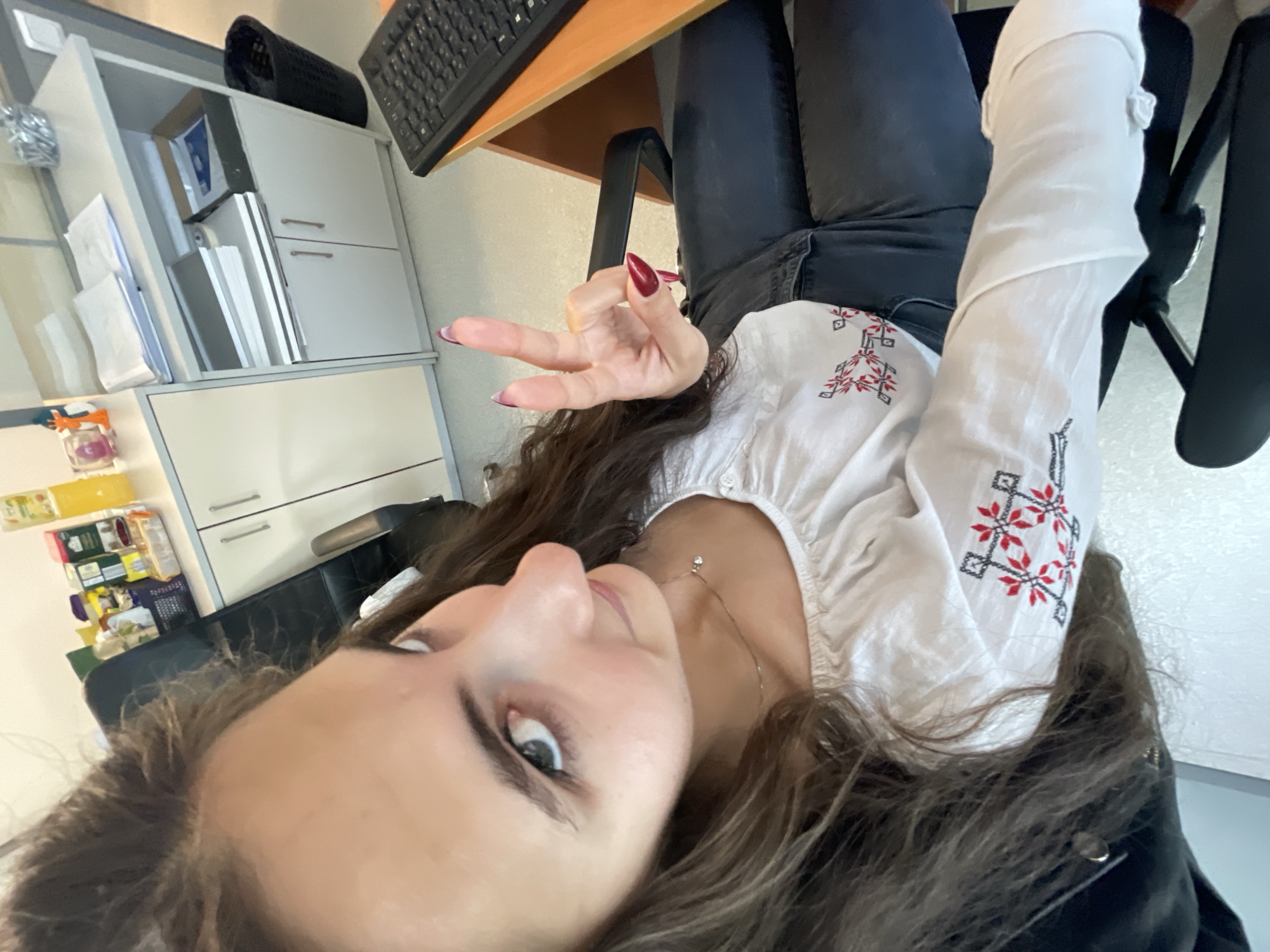 A woman is taking a selfie in what appears to be an office setting. She is sitting in a black office chair at a light-colored wooden desk with a black keyboard on it. She's wearing black jeans and a white blouse with red and black embroidery. Her hair is long and brown. She is holding up her hand, making a "peace" or "victory" sign. In the background, there is a white cabinet with drawers and shelves. The location is likely an office or study, possibly in Sofia, Bulgaria, but there is no specific indication within the image to confirm the Sofia location.