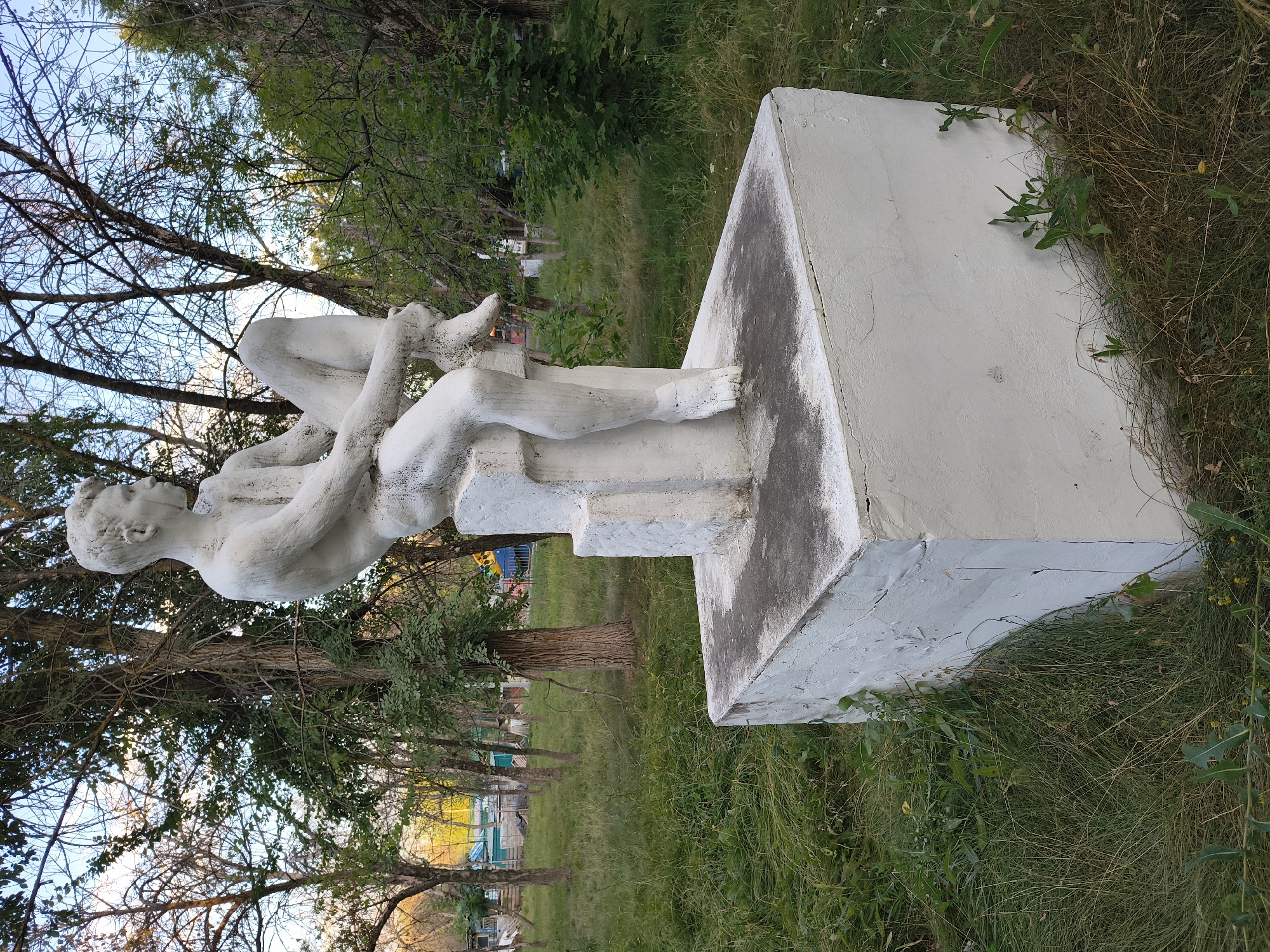 An outdoor shot captures a white concrete statue of a young man seated on a pedestal. The statue, positioned in the center of the frame, is rendered in a realistic style, with the figure depicted in a thoughtful pose, looking to the right. The sculpture is situated on a rectangular, tiered base, also made of white concrete, which is placed directly on the ground. The surrounding environment is a park-like setting with a variety of trees and overgrown green grass. In the background, through the trees, there are hints of structures and what appears to be a playground with colorful equipment, suggesting an urban recreational area. The lighting suggests it is daytime, possibly late afternoon or early morning given the quality of light and shadows. The overall scene implies a public space in Mikhaylovka, Russia, likely a park or a square.